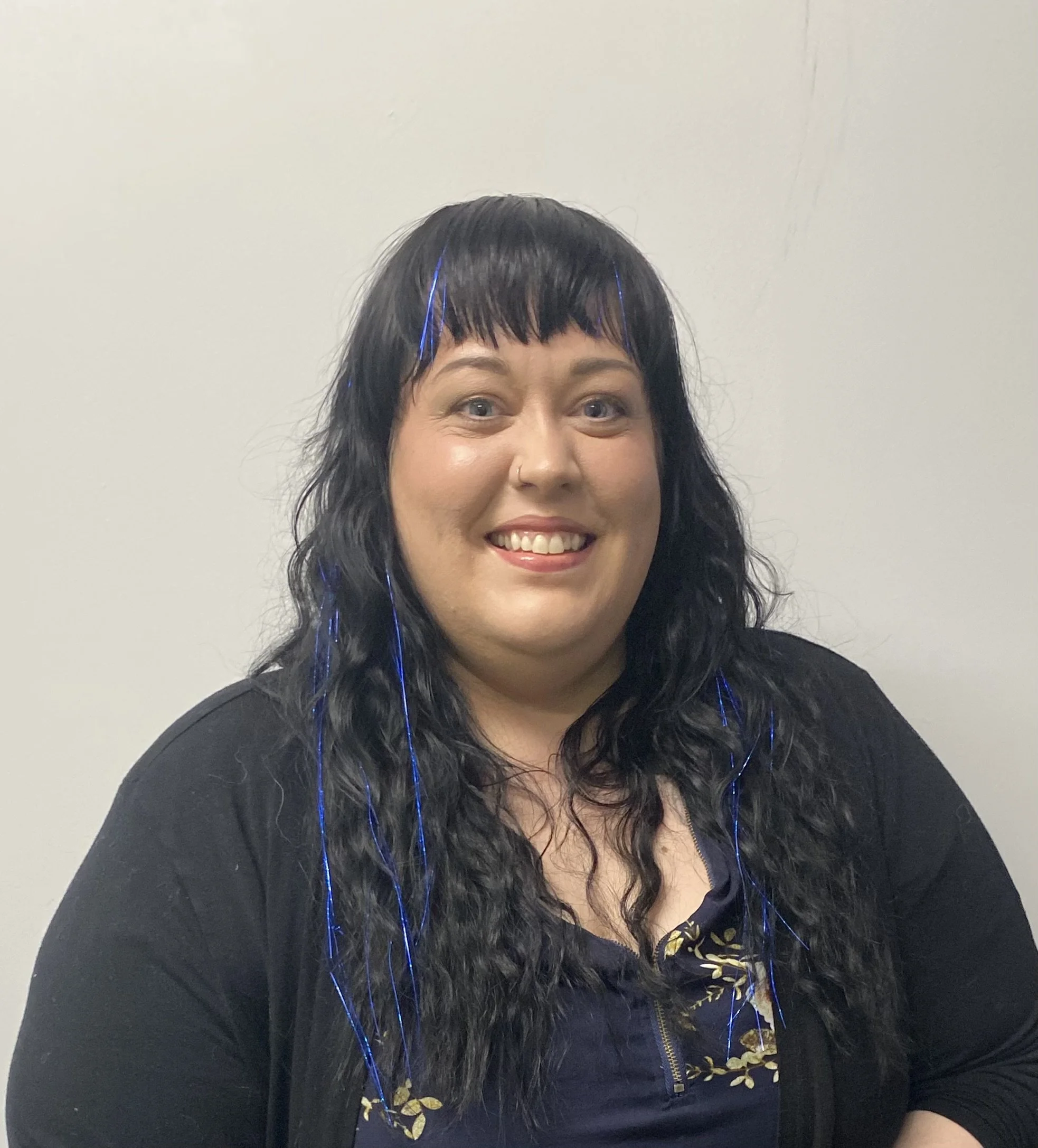 A woman with long, curly black hair and bangs, smiling, wearing a black top and blue shirt with floral details, standing against a plain white wall.