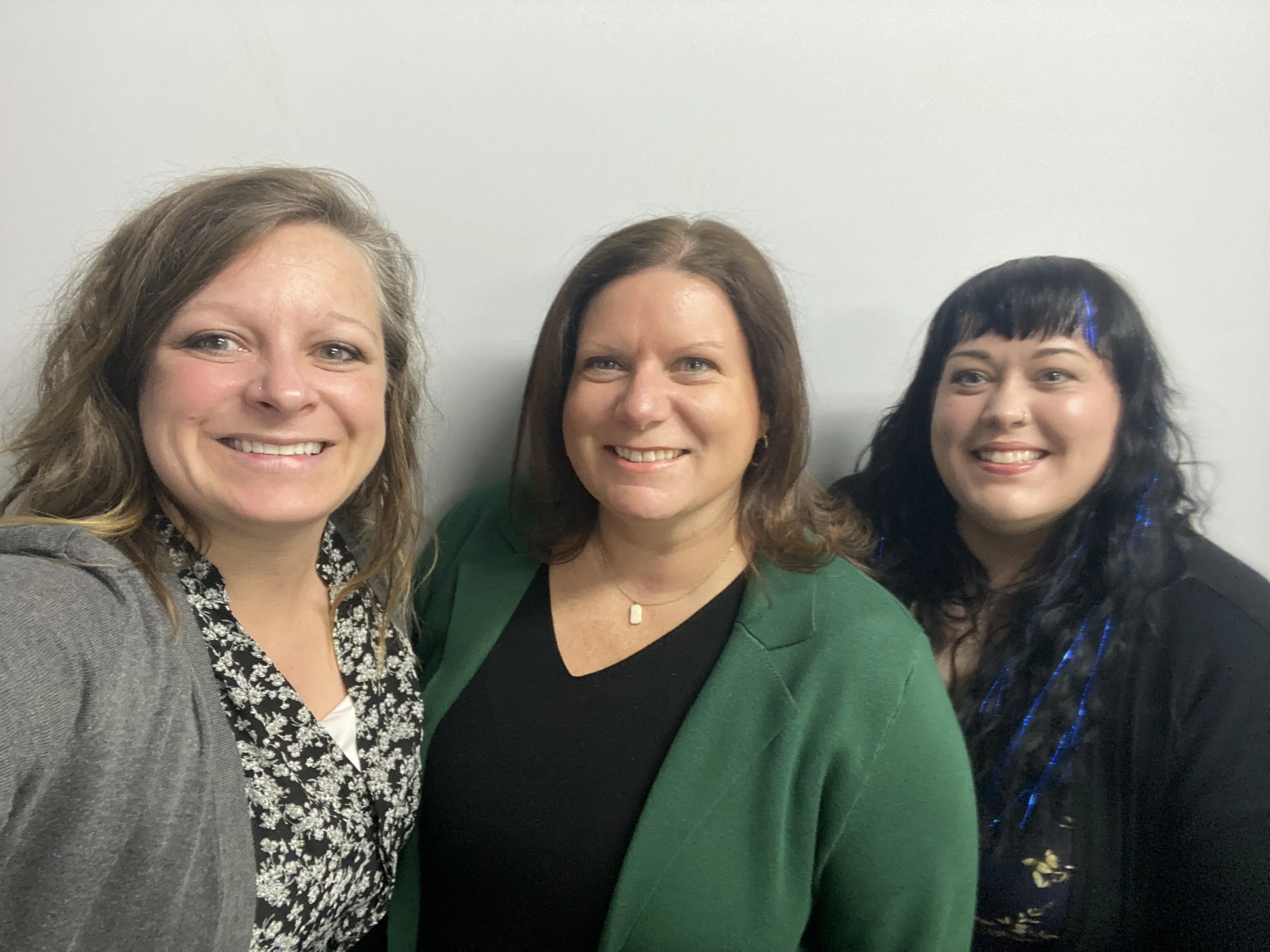 Three women smiling for a selfie, standing against a plain white wall. The woman on the left has wavy blonde hair and wears a grey blazer and patterned shirt. The woman in the middle has straight brown hair, wearing a black top and green blazer. The woman on the right has wavy black hair with blue streaks, wearing a black jacket over a floral dress.