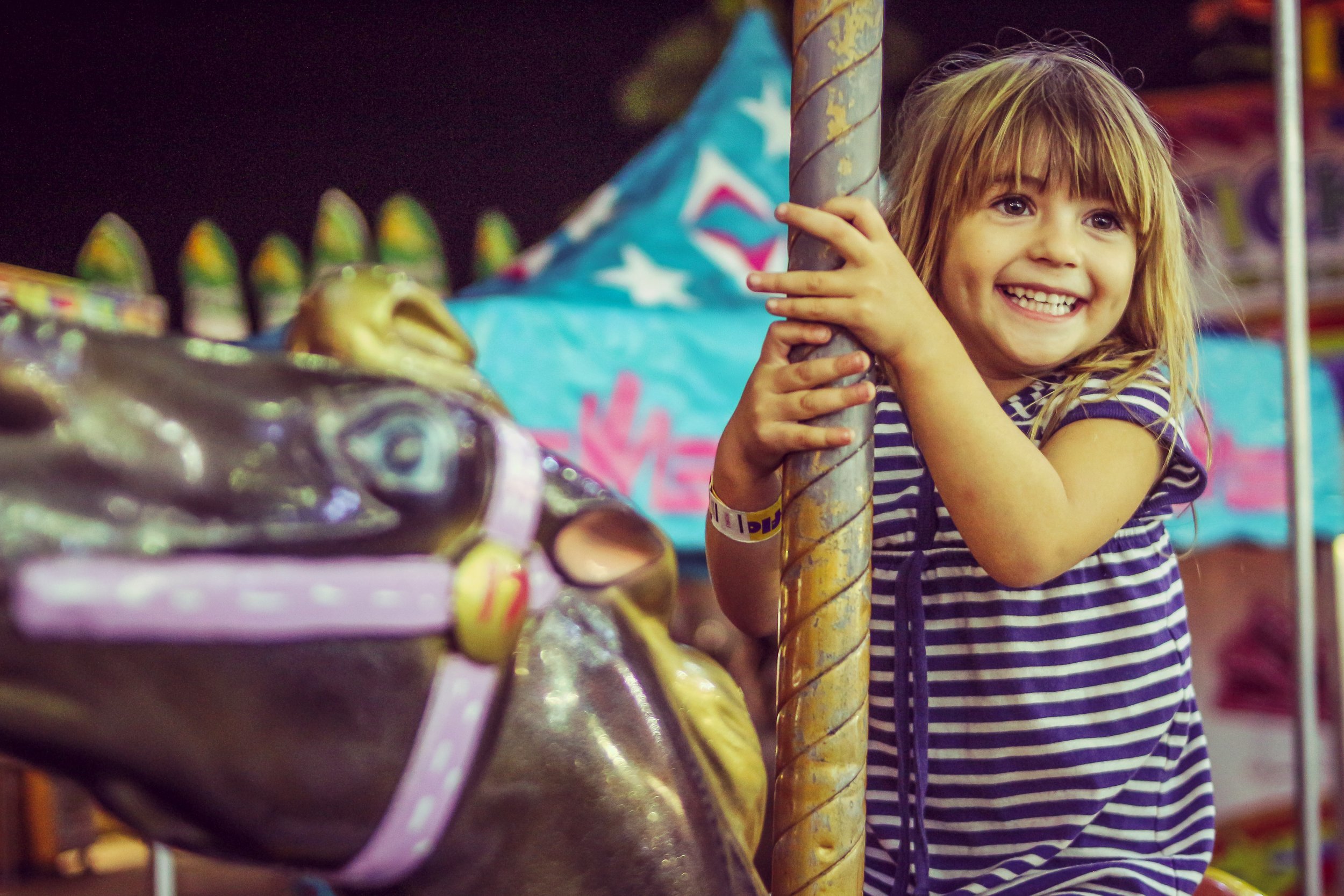 A young girl with long blonde hair and a striped dress is smiling and holding onto a carousel pole at a carnival or amusement park.
