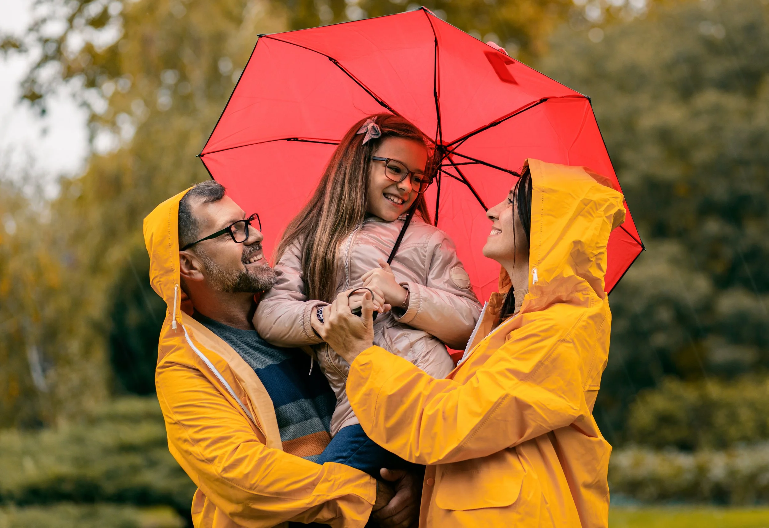 A family of three standing outdoors in the rain, holding a red umbrella. The father and mother, wearing yellow raincoats, are smiling as they hold their daughter up between them. The daughter is smiling and holding the umbrella handle, wearing glasses and a pink jacket.