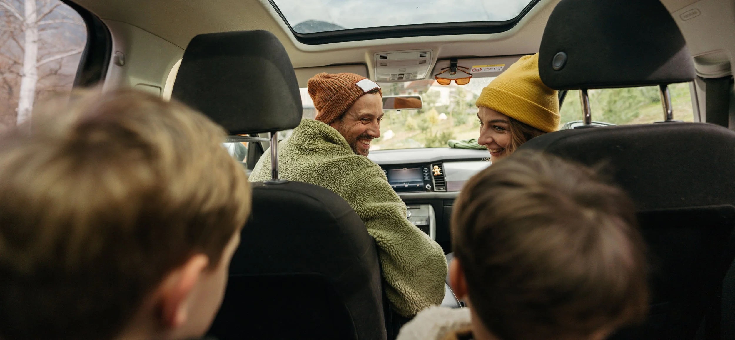 A group of people sitting in the backseat of a car, smiling and talking to each other. Two adults are visible, one man and one woman, both wearing beanies, with children in the foreground.