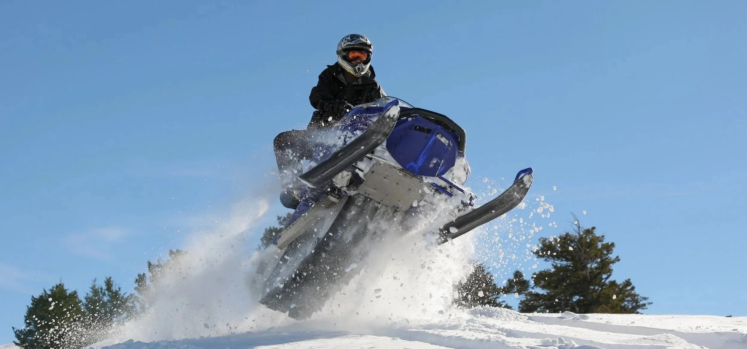 A person riding a snowmobile in snowy terrain with pine trees in the background under a clear blue sky.