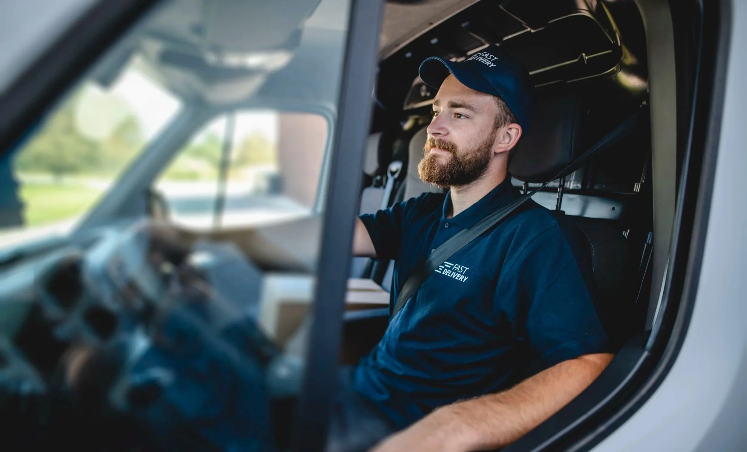 A delivery driver with a beard and a blue hat and shirt sitting in the driver's seat of a delivery truck, looking out the window.