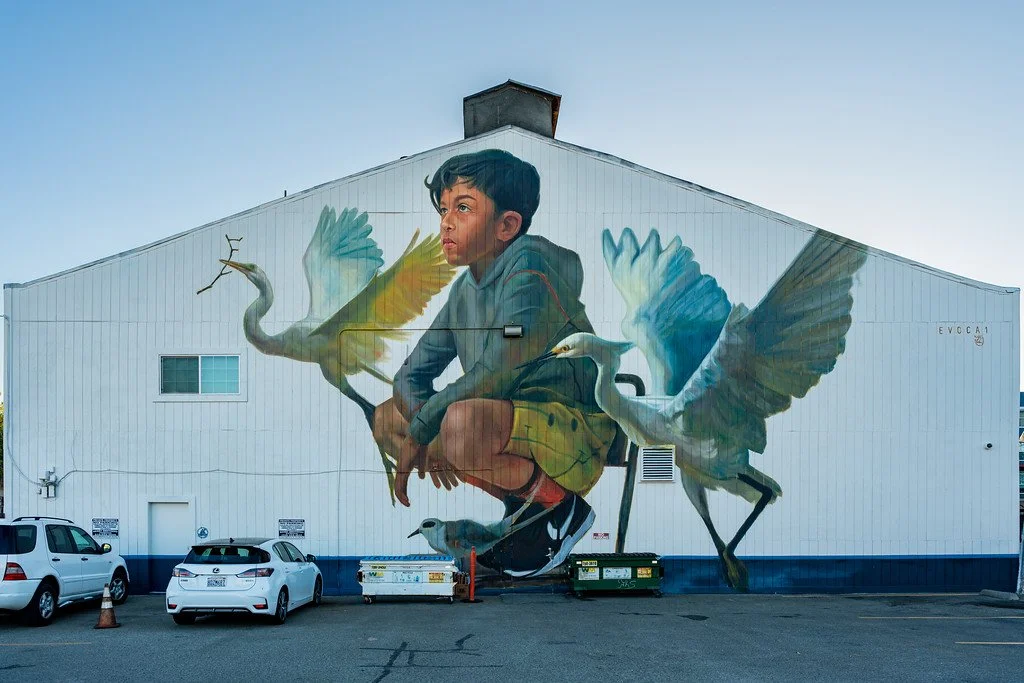 A mural of a young boy squatting surrounded by four large flying cranes painted on the side of a building.