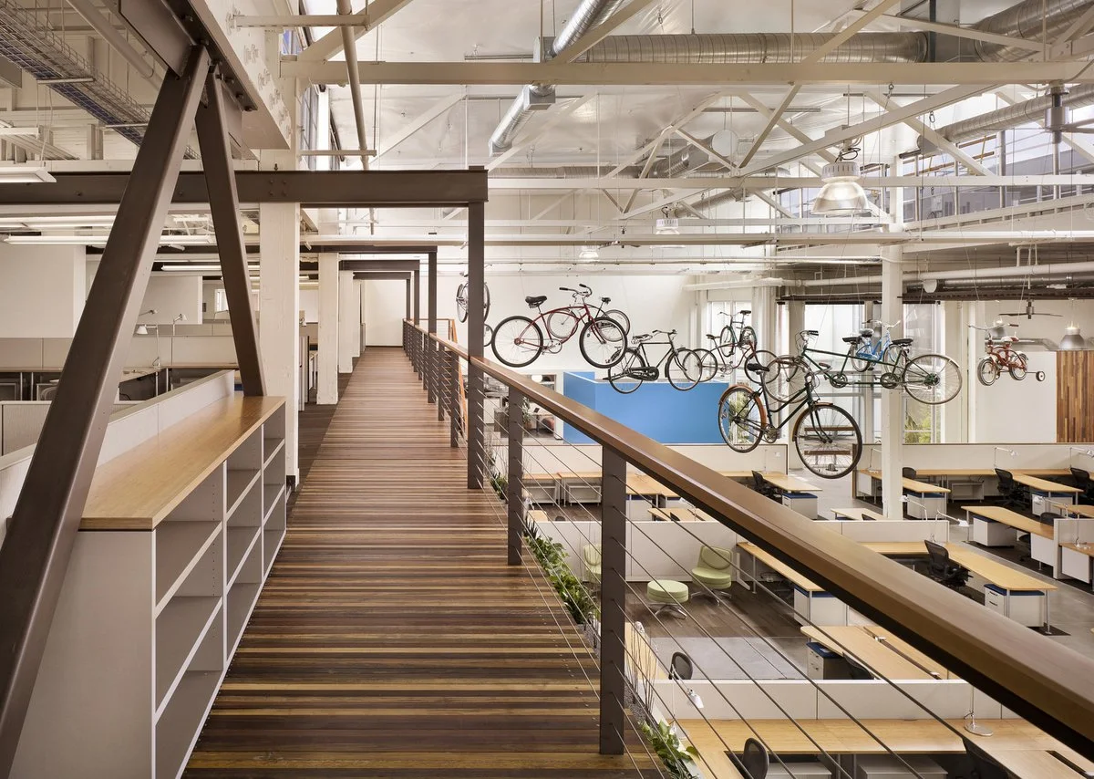 An office interior with a second-floor walkway overlooking work stations below. Bicycles are hanging from the ceiling, and there are desks with chairs, computers, and green sitting areas.