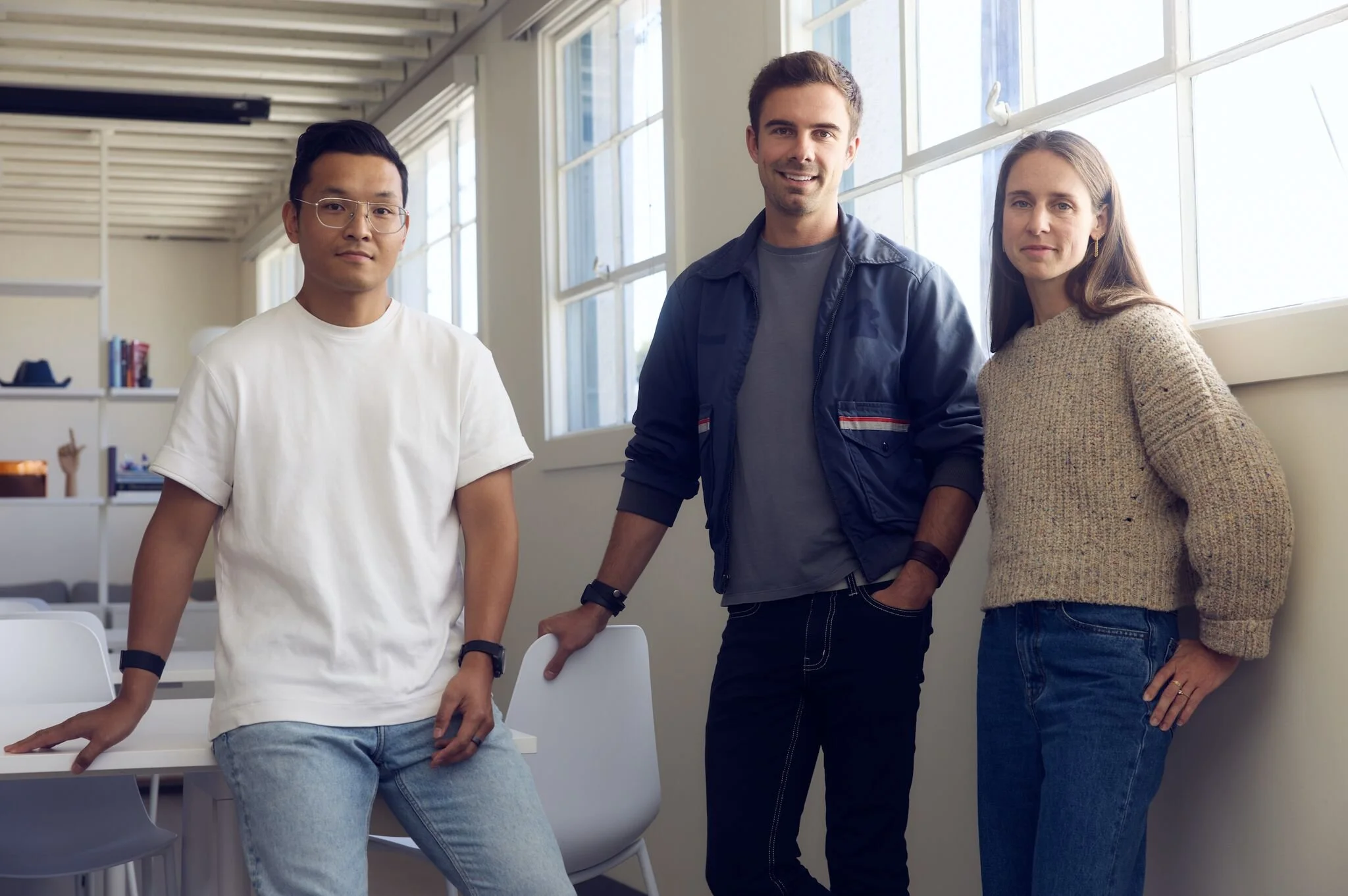 Three young adults standing together in a modern classroom with large windows. They are smiling and casually dressed, with one leaning on a chair.