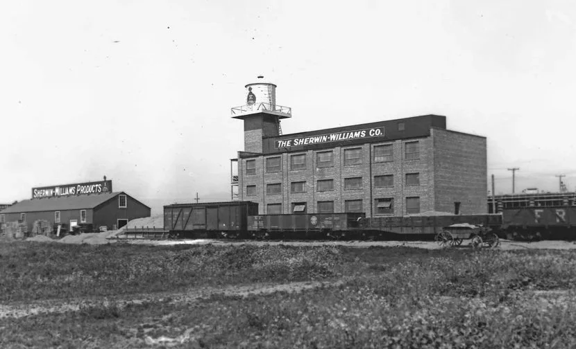Black and white photo of an old industrial building labeled 'The Sherwin-Williams Co.' with a water tower on top, a smaller building in the background labeled 'Sherwin-Williams Products,' and train cars on tracks in the foreground.