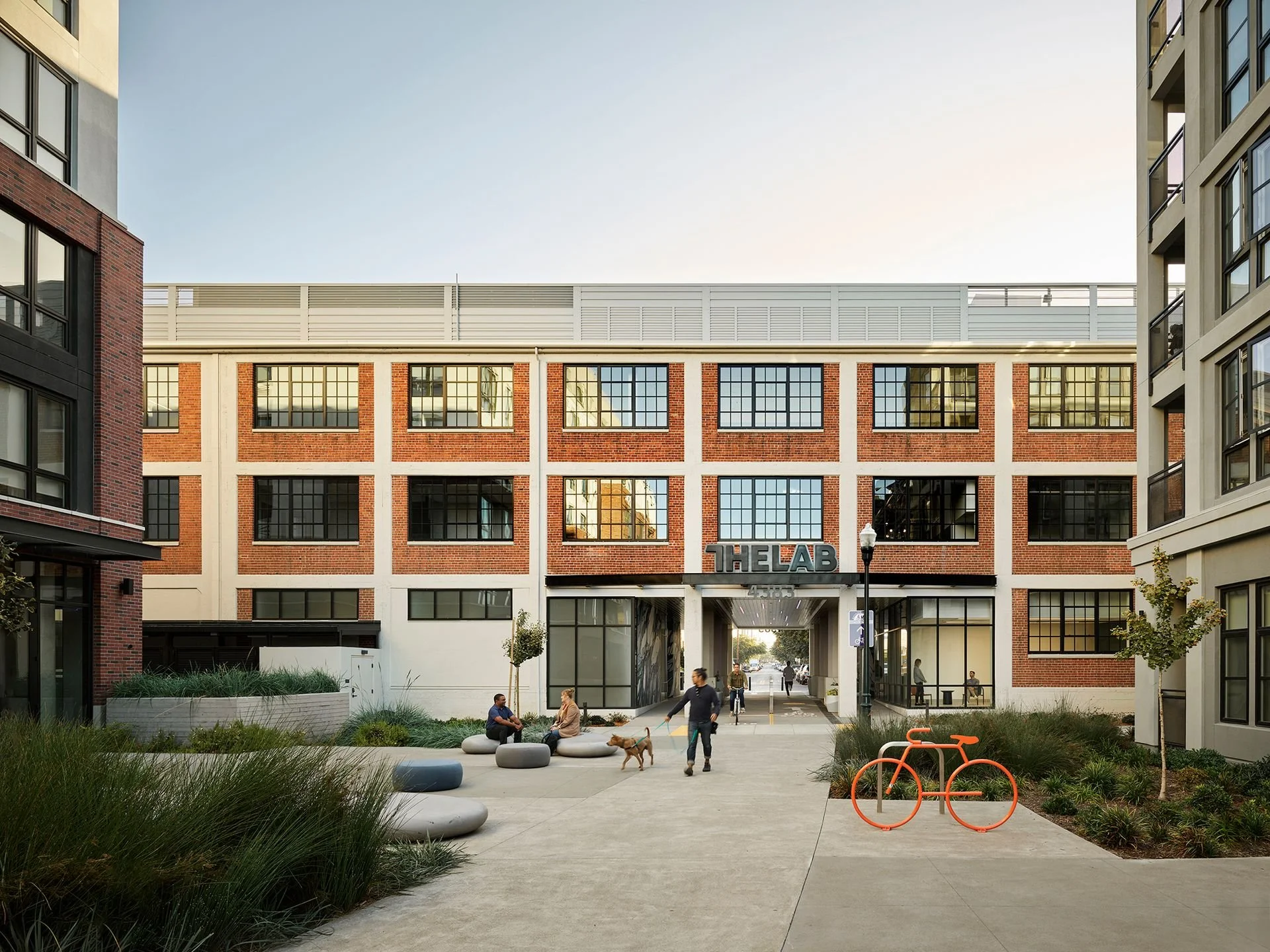A modern, red-brick building with large glass windows and a sign that reads THE LAB. There is a small park in front of the building with people sitting on concrete seats, walking, and a dog. An orange bike rack is also visible.