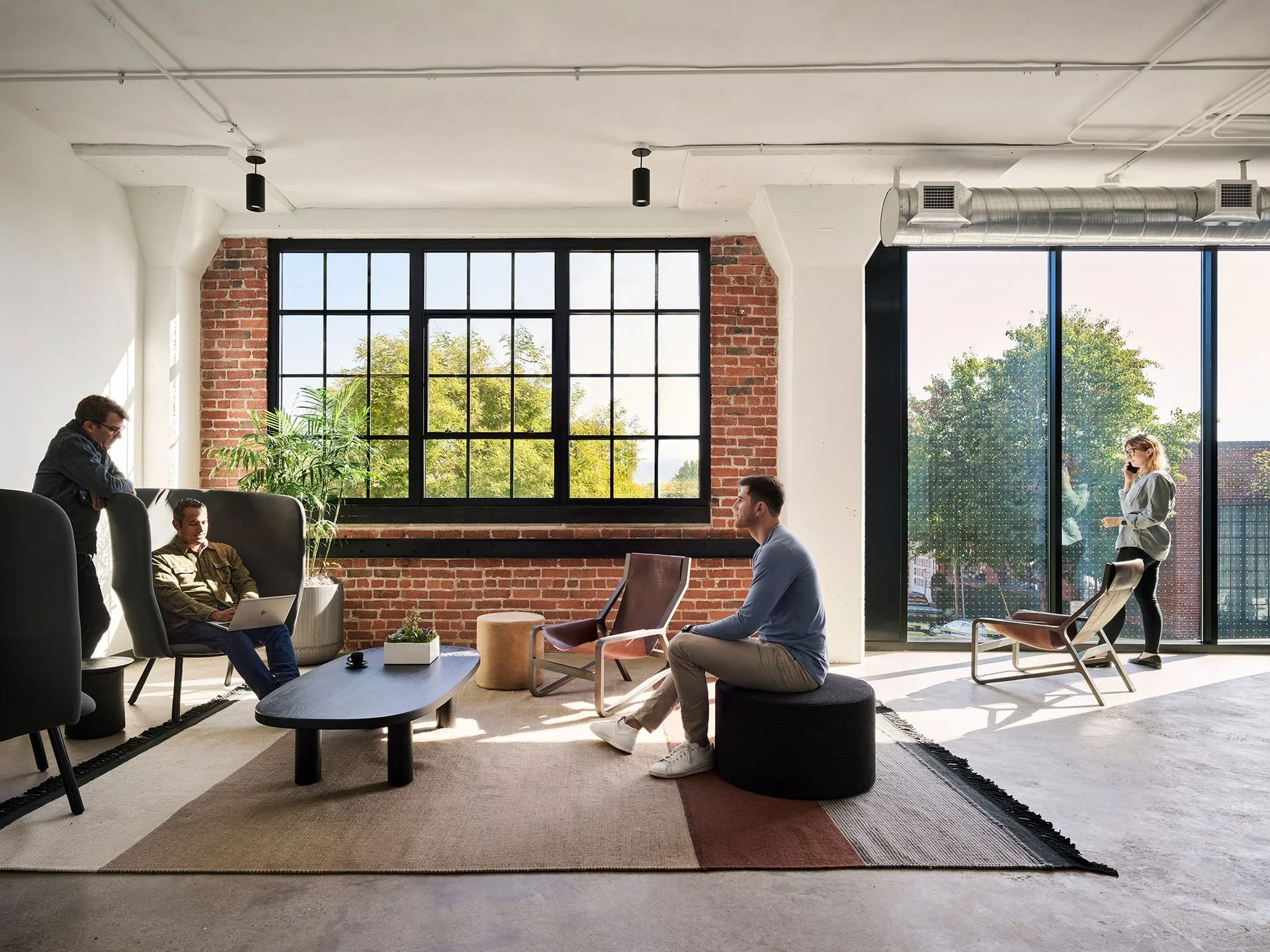 Modern office space with large windows, exposed brick walls, and several people sitting and talking, some working on laptops, with indoor plants and contemporary furniture.