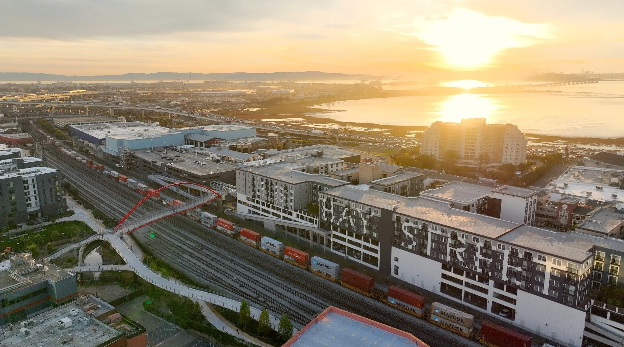 A cityscape during sunset featuring buildings, roads, and a body of water in the background.