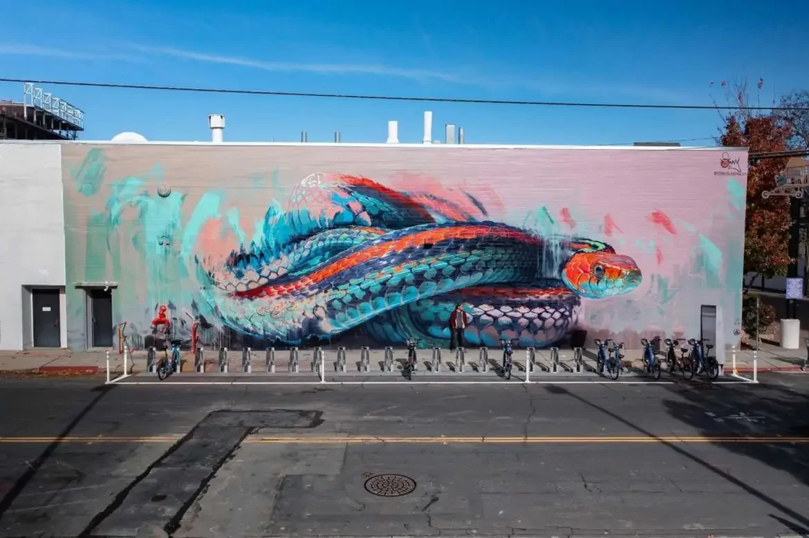 Colorful street mural of a large snake with vibrant blue, orange, and black scales on a pink background, painted on the side of a building.