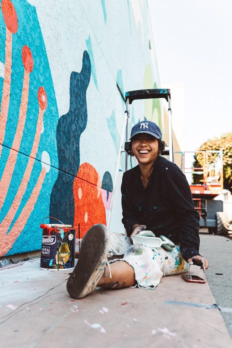 A woman with a New York Yankees cap sitting on the ground, painting a colorful mural on a wall. She has a big smile and is wearing paint-splattered clothes. There are painting supplies nearby and a lift in the background.