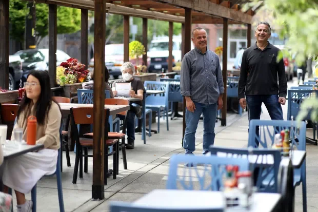 A busy outdoor restaurant patio with several people dining and two men standing and chatting, with other diners seated at tables.