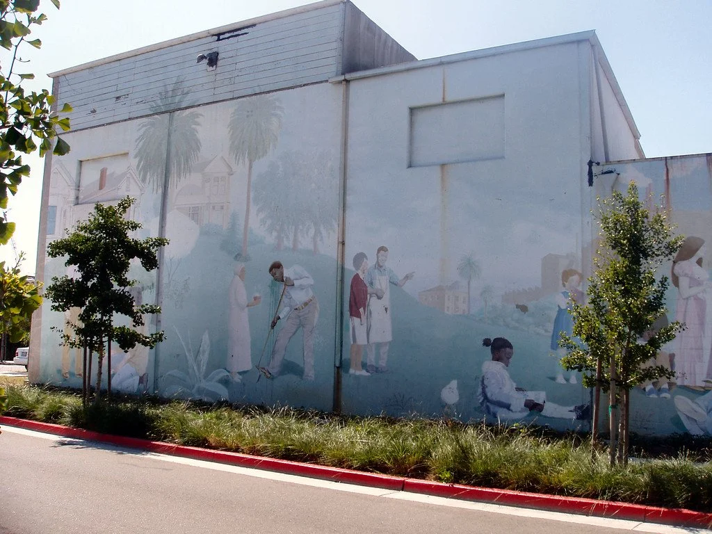A mural on a building depicts medical professionals and patients in a healthcare setting, with trees in the foreground and a cityscape background.