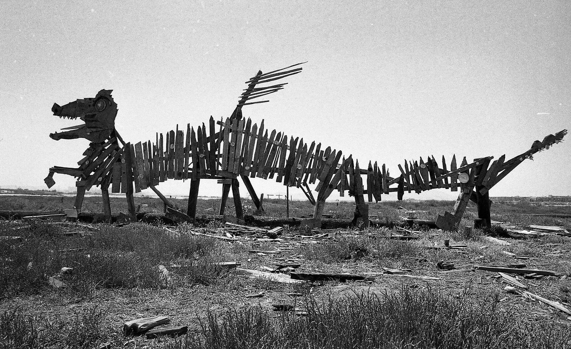A large sculpture of a dinosaur made from wooden planks and metal parts, located outdoors in a field with grass and scattered debris, under a cloudy sky.