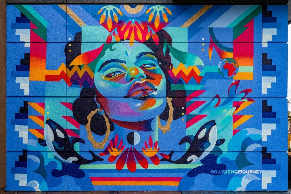 Colorful mural of a confident woman with curly hair, large earrings, and vibrant clothing, surrounded by abstract patterns, bright colors, and a small depiction of the Earth.