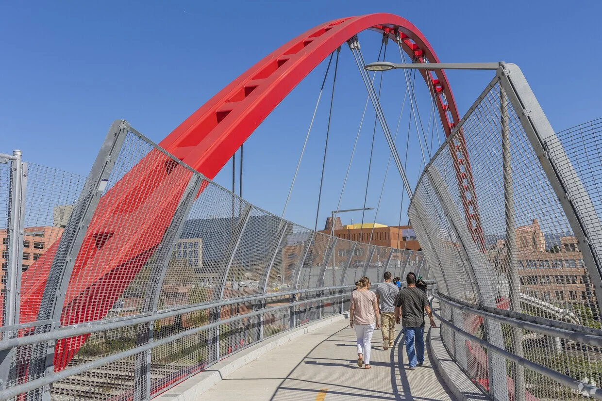 People walking on a modern, curved pedestrian bridge with a red arch and metal fencing, with city buildings in the background.