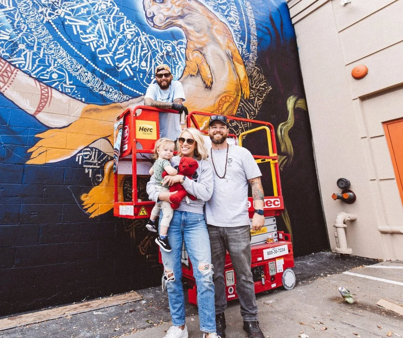 A group of four people standing in front of a colorful mural on a brick wall. The mural depicts a large animal, possibly a lion, with intricate designs and a blue mane. The group includes a woman holding a child holding a red teddy bear, a man with tattoos, and another man standing in a scissor lift. They are smiling and appear to be enjoying their time together.