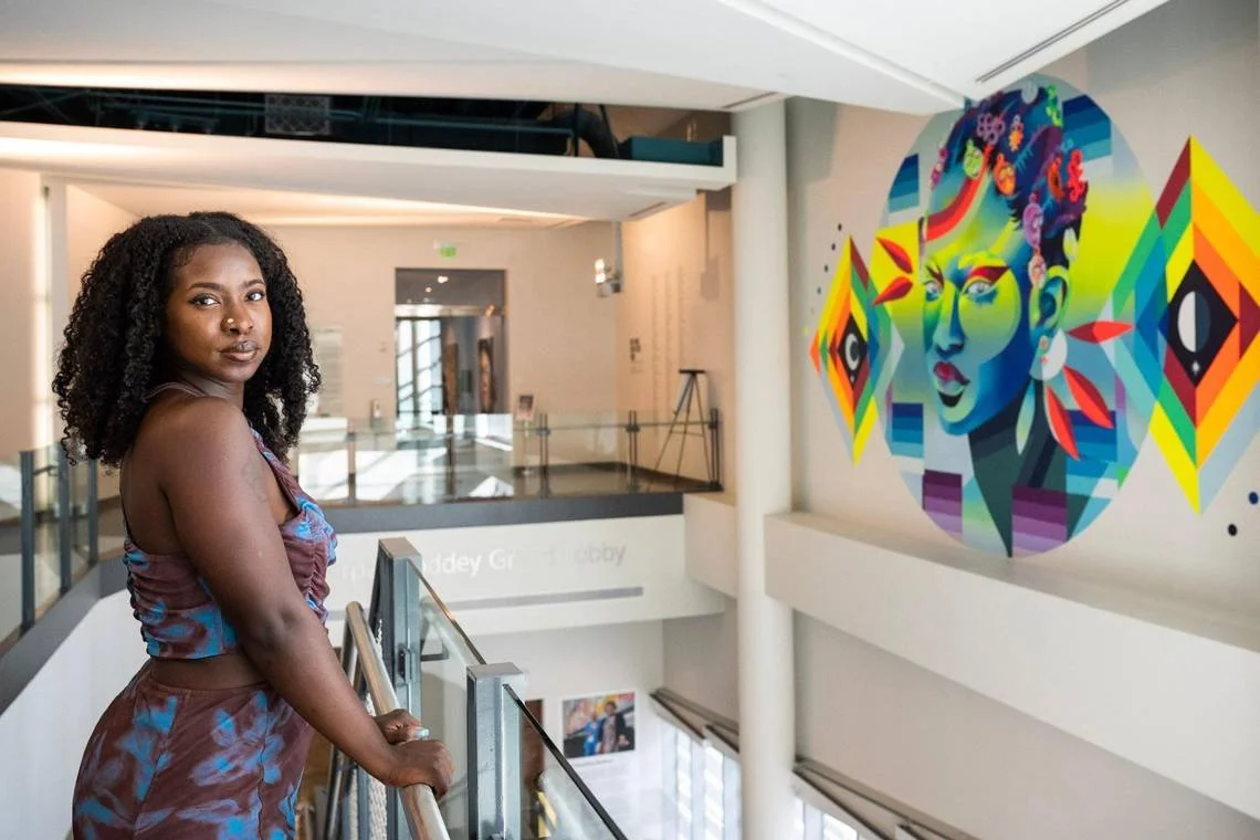 A woman with curly hair in a purple and blue outfit stands on an indoor balcony of an art gallery, looking at the camera. Behind her is a colorful portrait mural of a woman with vibrant hair and geometric floral designs.