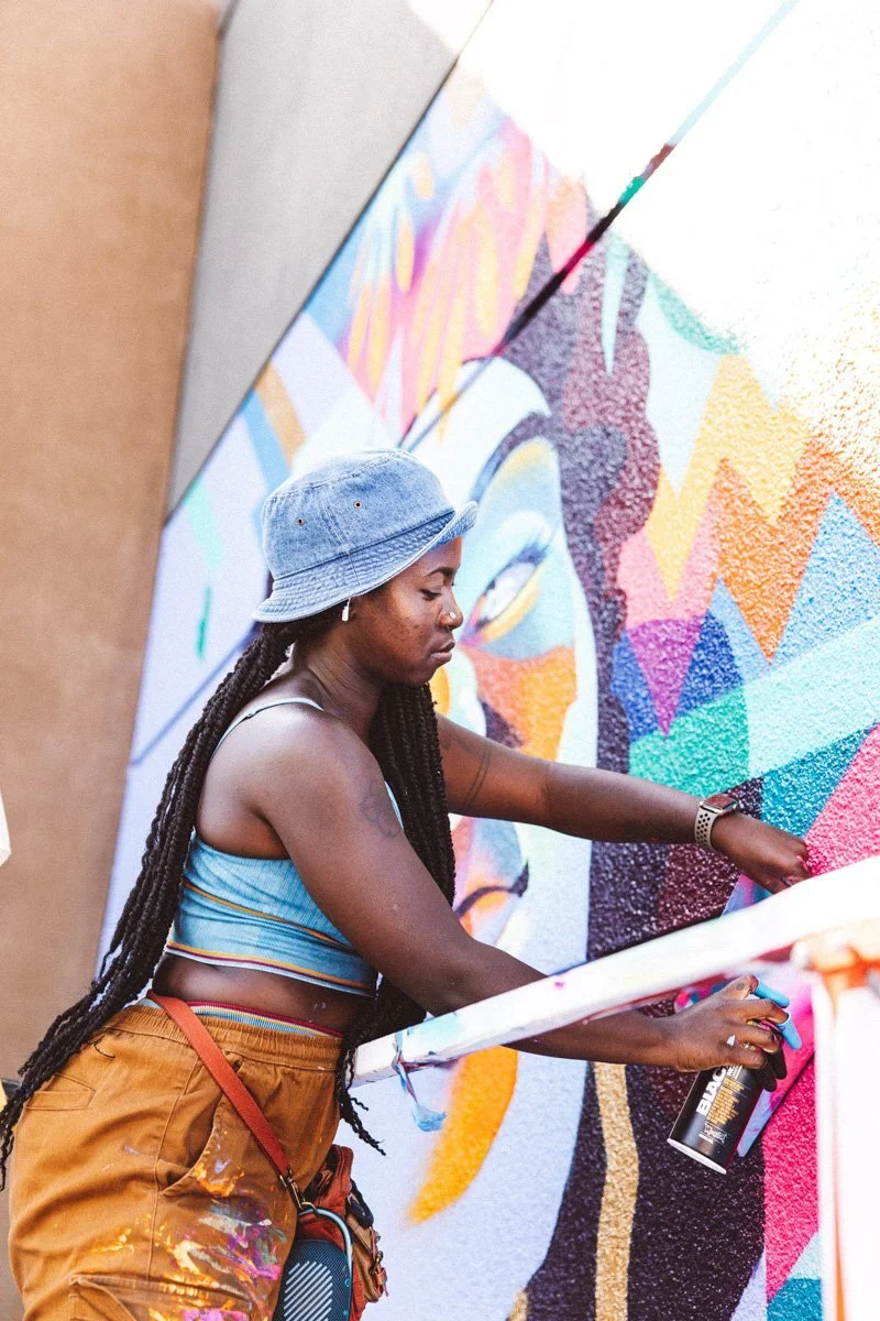 A woman with long braided hair, wearing a blue bucket hat, a striped crop top, and orange paint-splattered shorts, spray-painting a colorful mural on a wall.