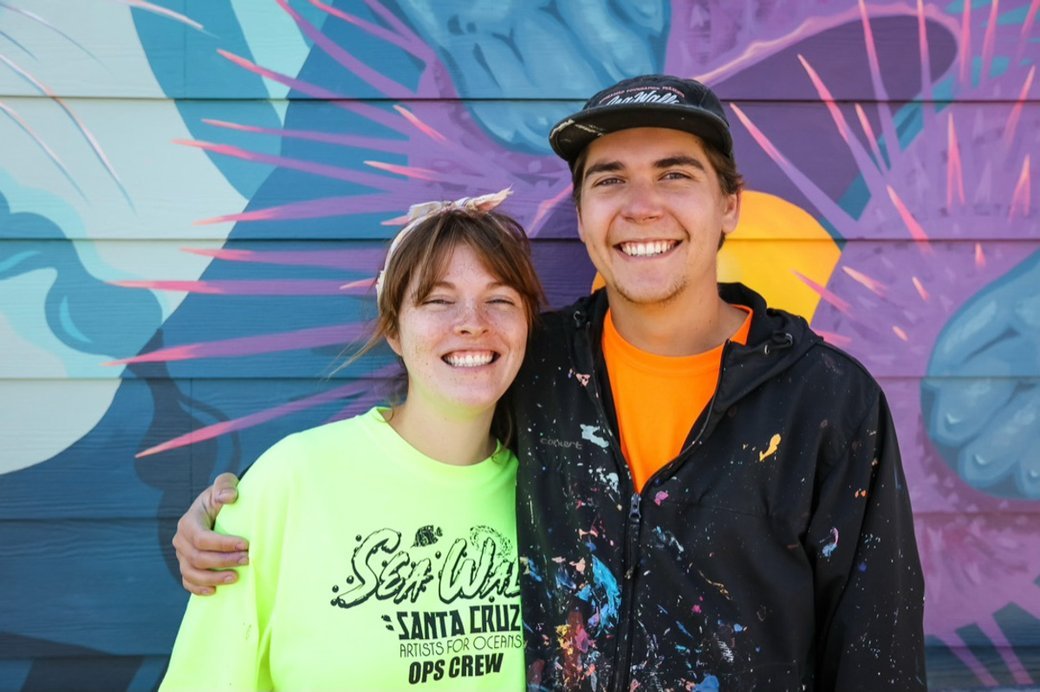 Two young smiling people standing in front of a vibrant graffiti mural, one wearing a neon yellow shirt with green and black text, the other in a black jacket with paint splatters, posing with their arms around each other.