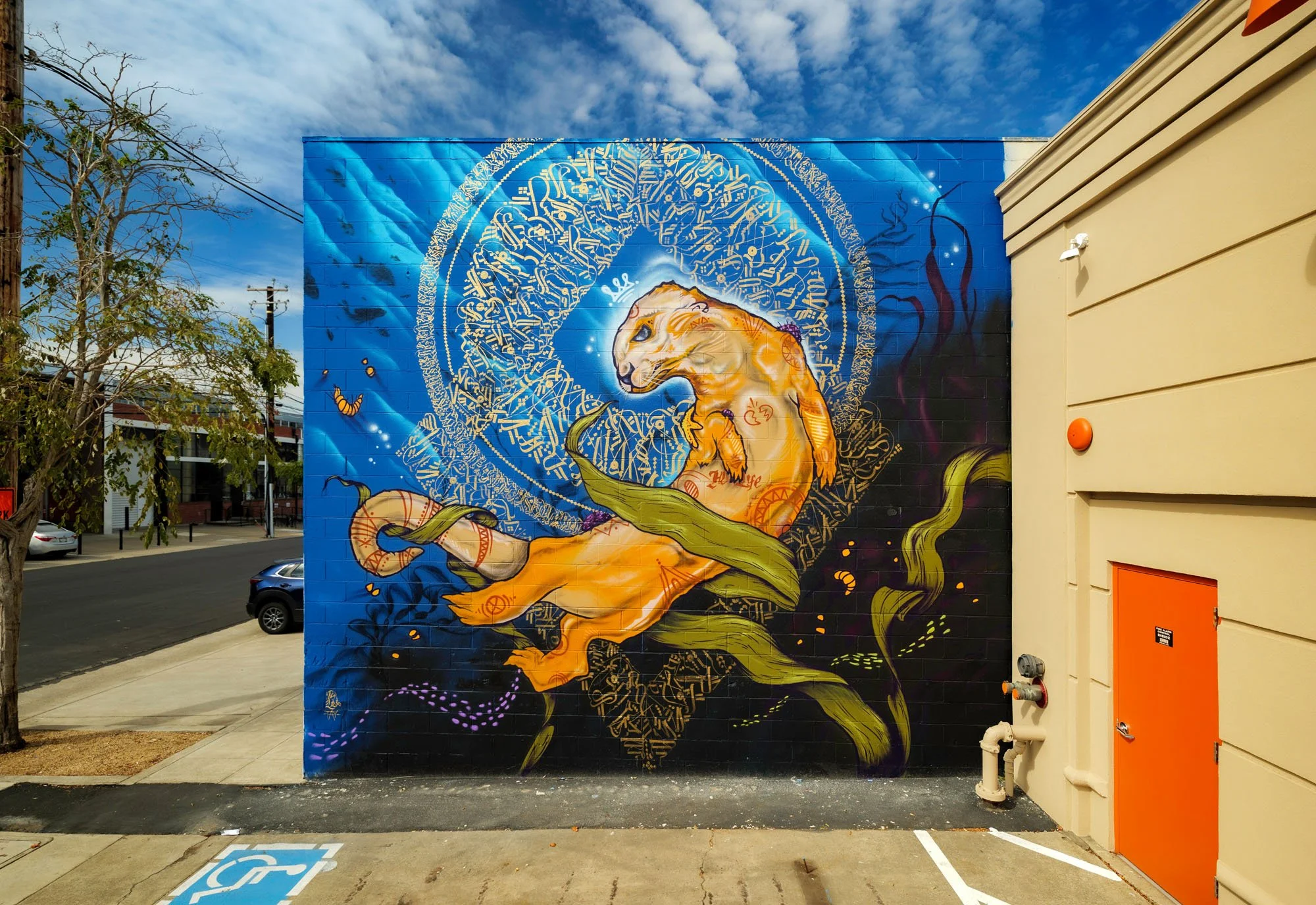 Colorful mural of a feline figure with intricate gold and blue designs on a bright blue background, painted on an exterior wall.