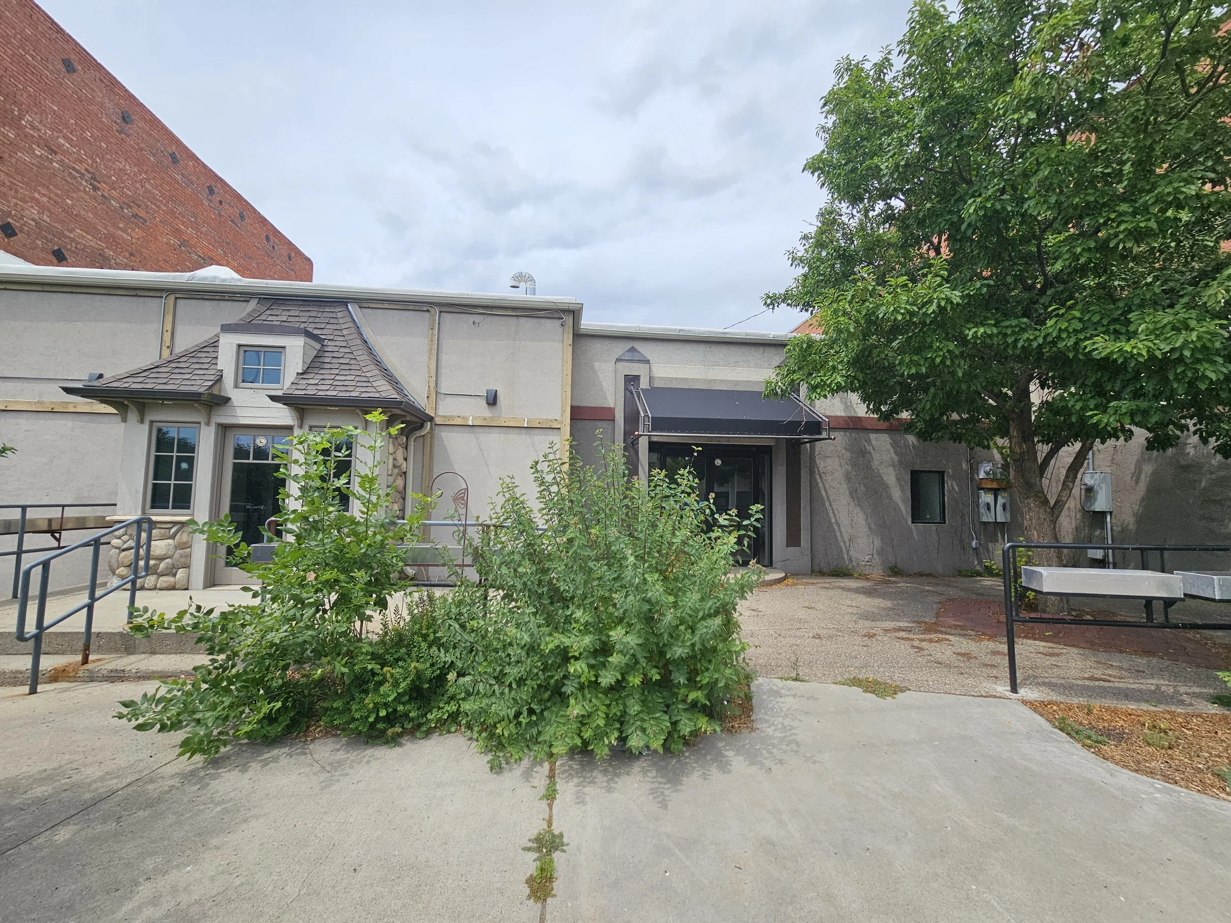 An outdoor scene with a small building that has two windows and a door, surrounded by greenery including a large tree on the right, and a paved pathway and some small plants in the foreground. The sky is overcast.