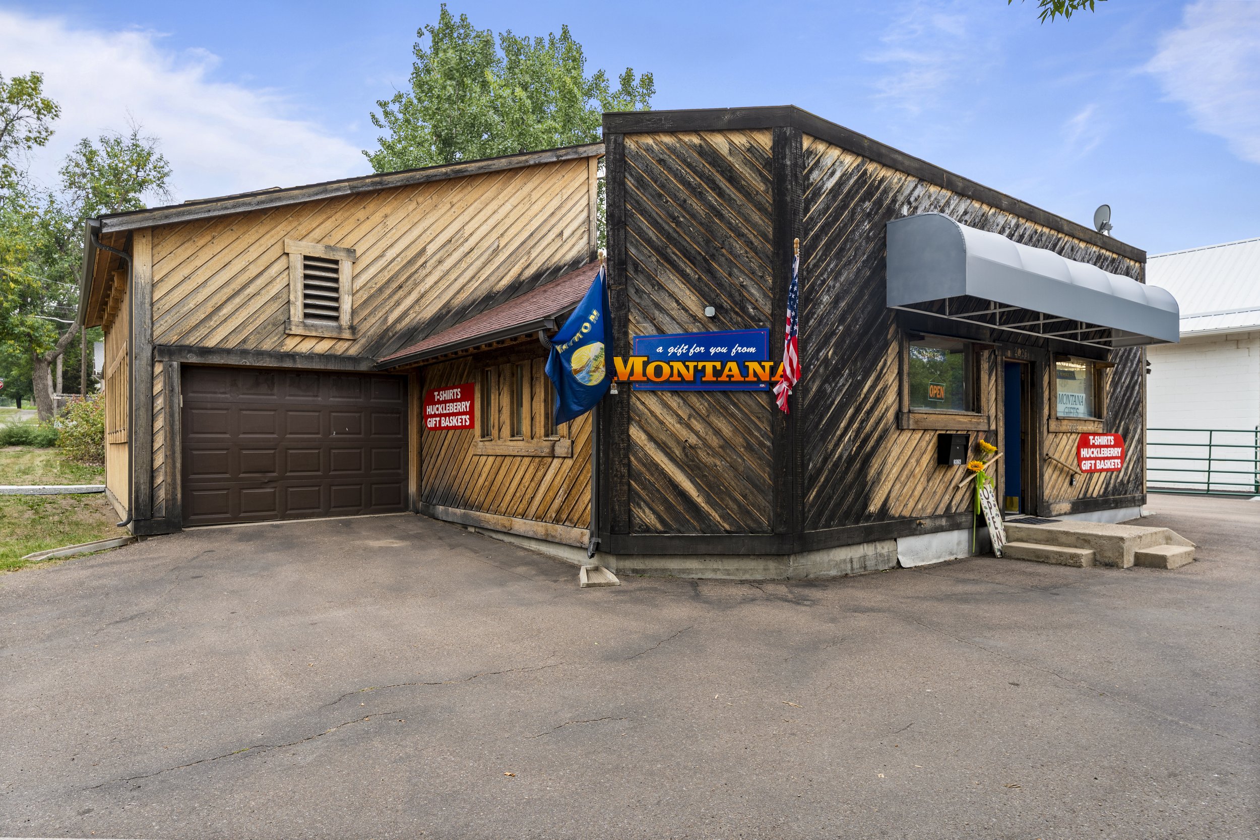 Front of a small store with a wooden exterior, signs advertising T-shirts, huckleberry gift baskets, and a Montana gift shop, along with flags and a sign that says 'a gift for you from Montana'.