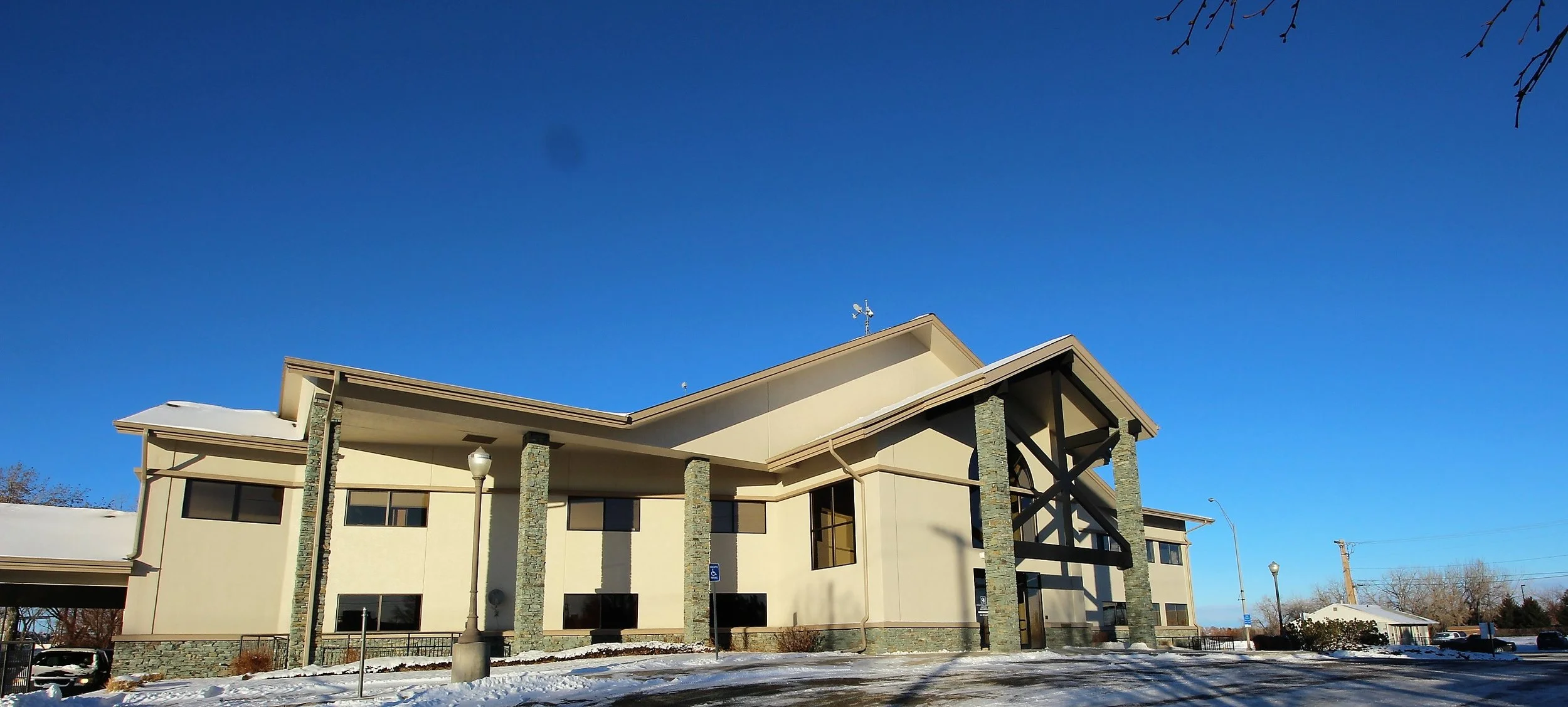 A modern two-story building with beige walls, stone pillars, and large black-framed windows, under a clear blue sky with some snow on the ground nearby.