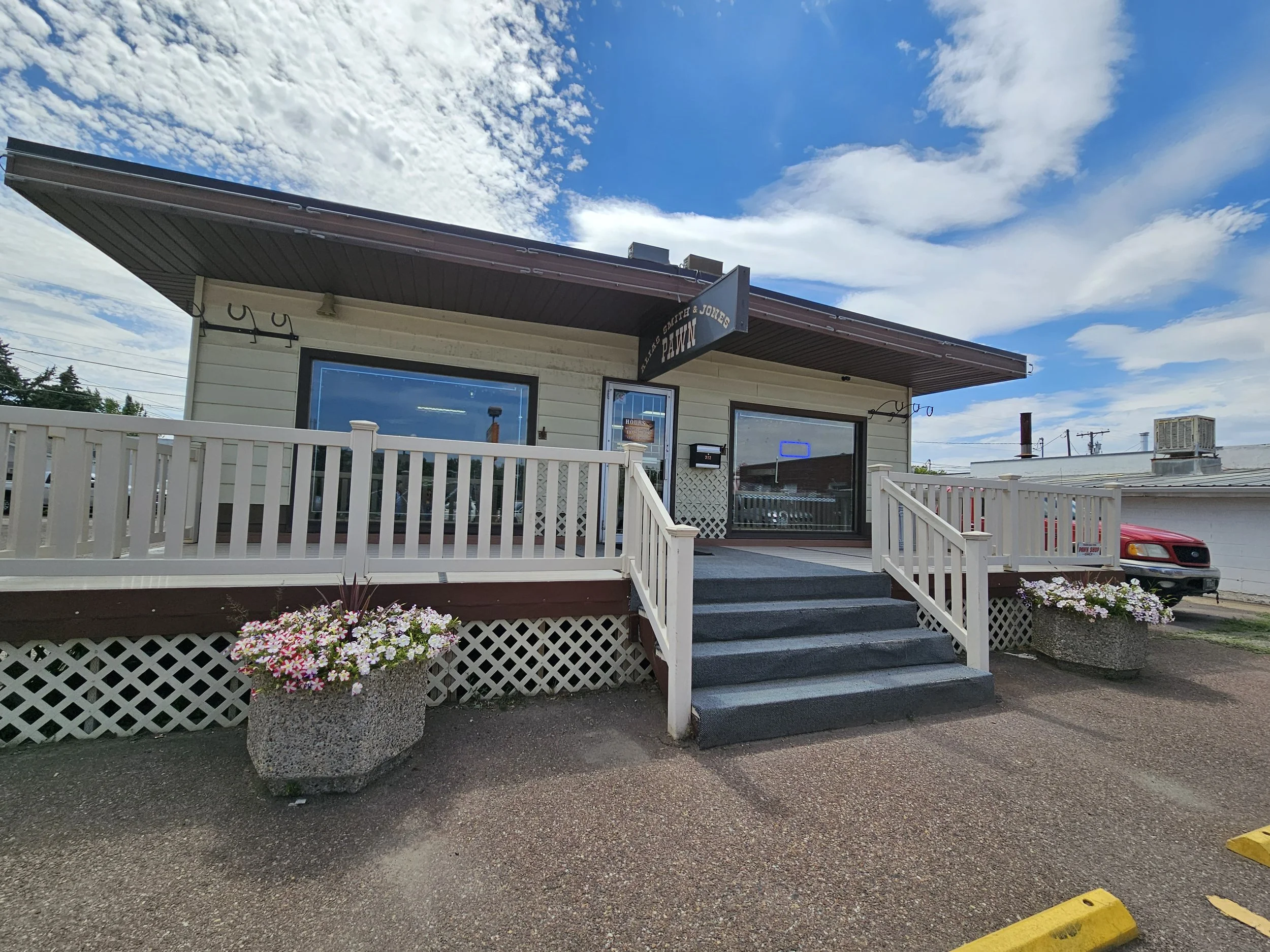A small pawn shop with a sign that reads 'Smith & Jones Pawn' on the front of the building. The building has a porch with a white railing and a vending machine near the entrance. There is a large potted plant with pink and white flowers in front of the porch. The sky is partly cloudy with patches of blue visible.