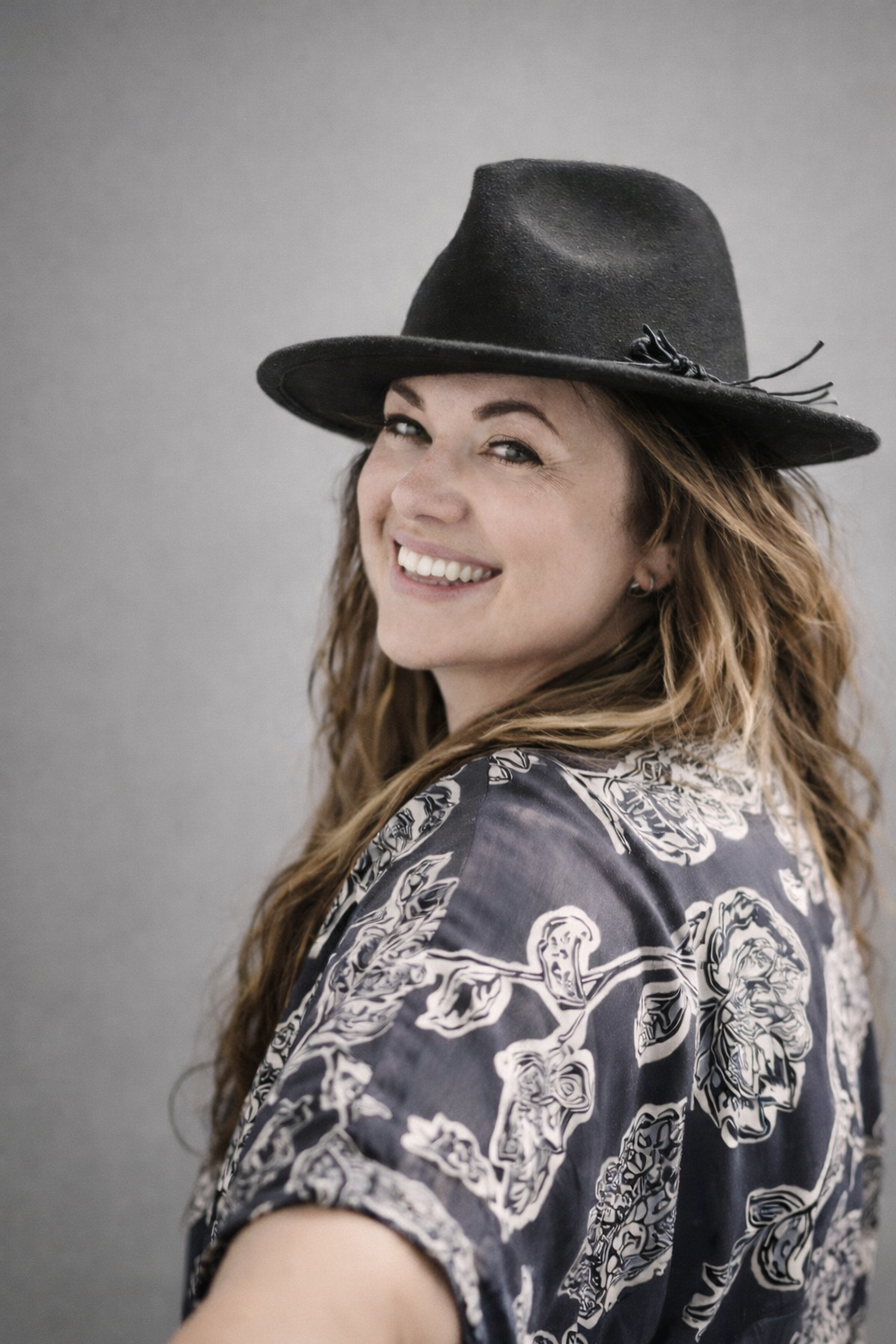 A woman smiling, wearing a black hat and a patterned shirt, standing against a plain gray background.
