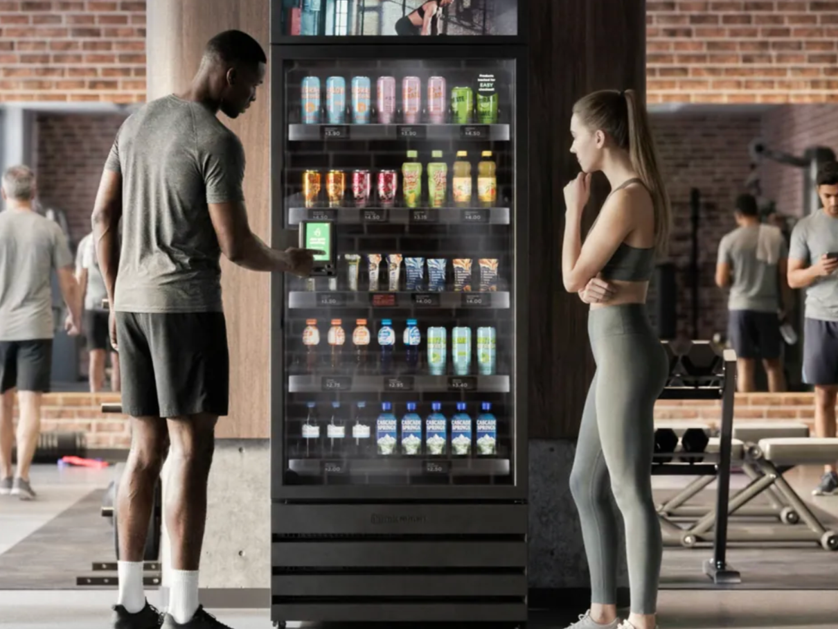A man and a woman standing in front of a vending machine in a gym. The man is using his phone to make a selection, while the woman looks on. There are other gym members in the background.