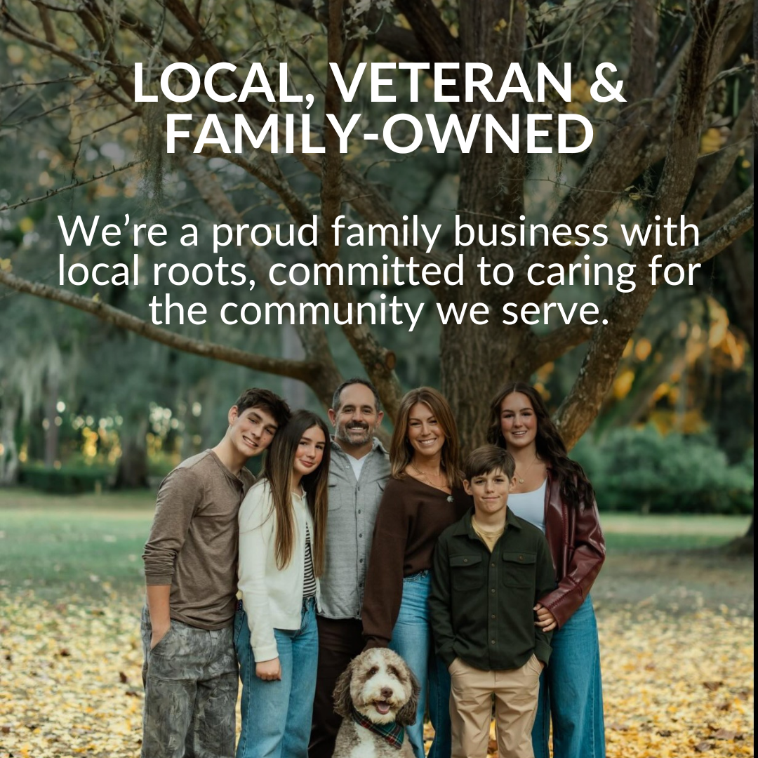 A family with three adults, two teenagers, a young boy, and a dog, standing outdoors under a large tree, smiling.