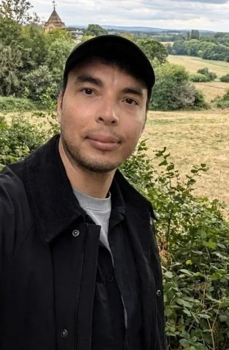 A man taking a selfie outdoors with greenery and fields in the background, a church steeple visible in the distance, overcast sky.