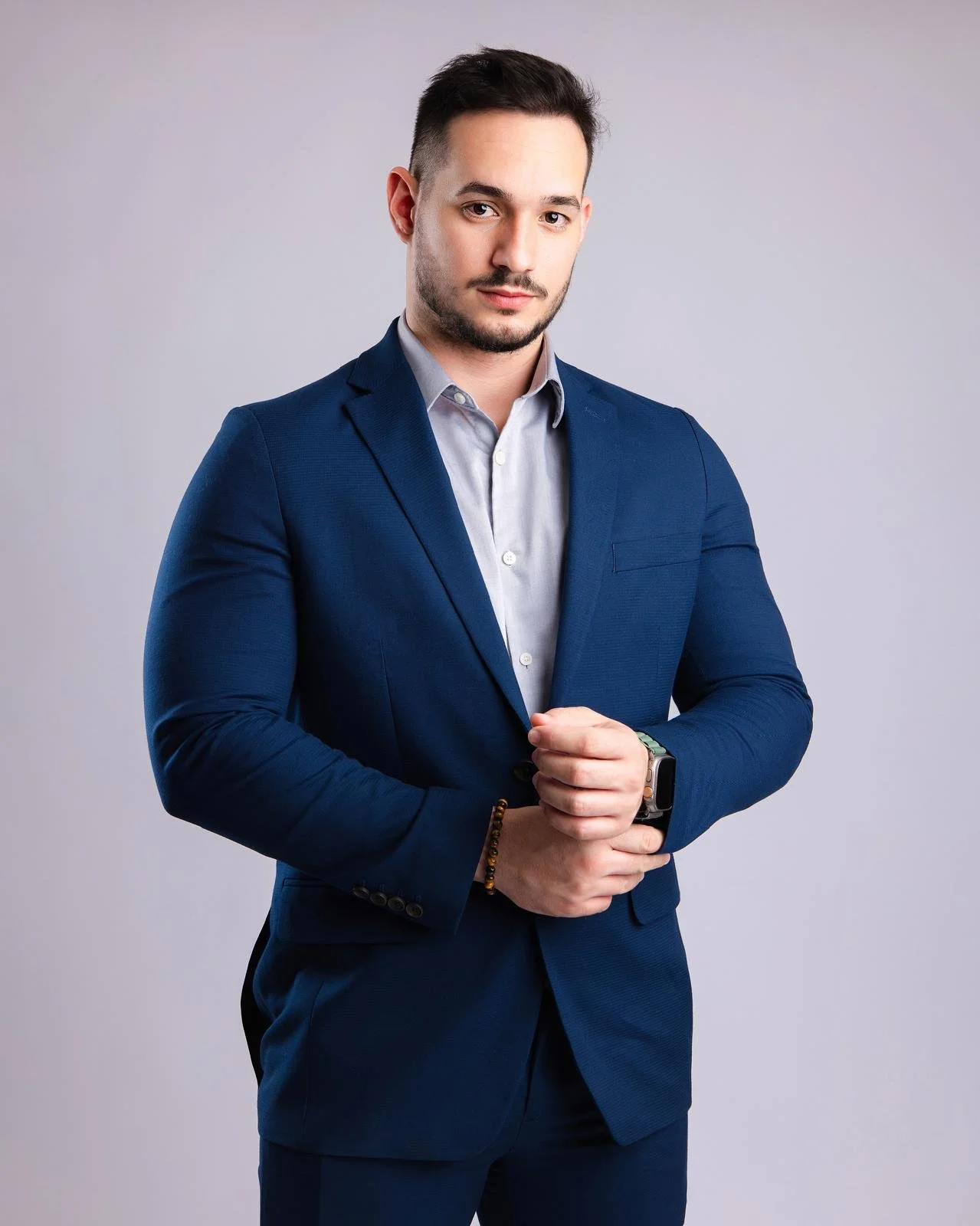 Hombre en traje azul y camisa gris, posando con confianza, con fondo gris claro.