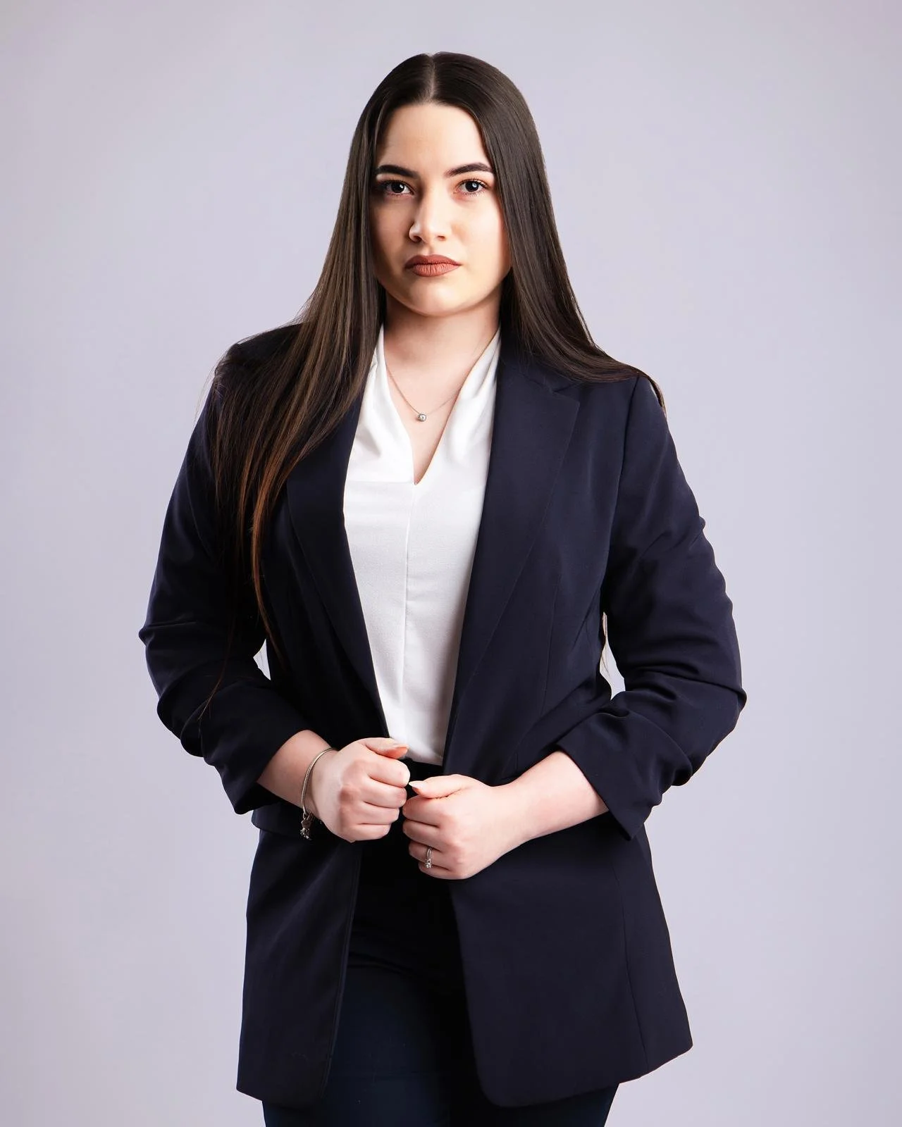 Mujer joven con cabello largo y oscuro, vestida con blazer negro y blusa blanca, en un fondo gris claro.