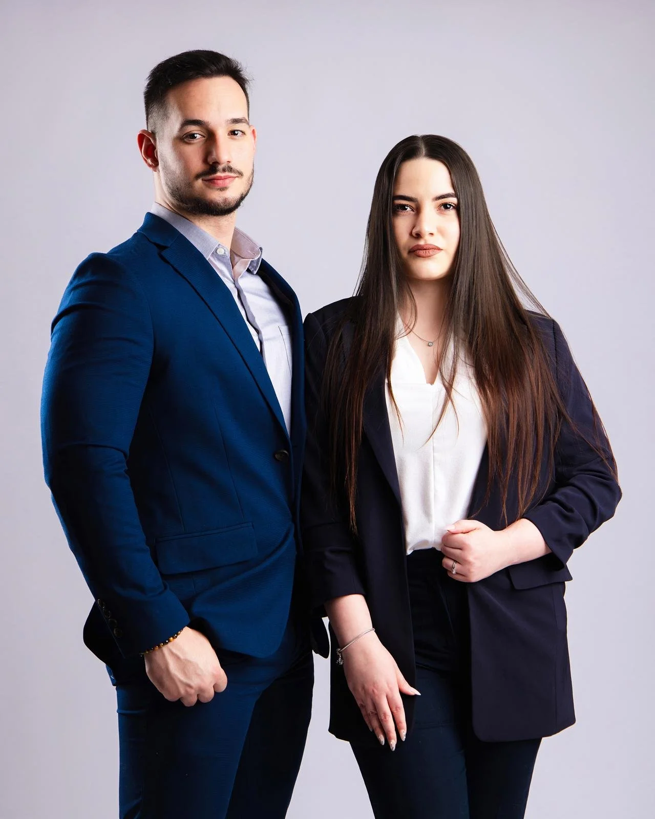 Un hombre y una mujer de negocios posando juntos, ambos con ropa formal y frente a un fondo gris.
