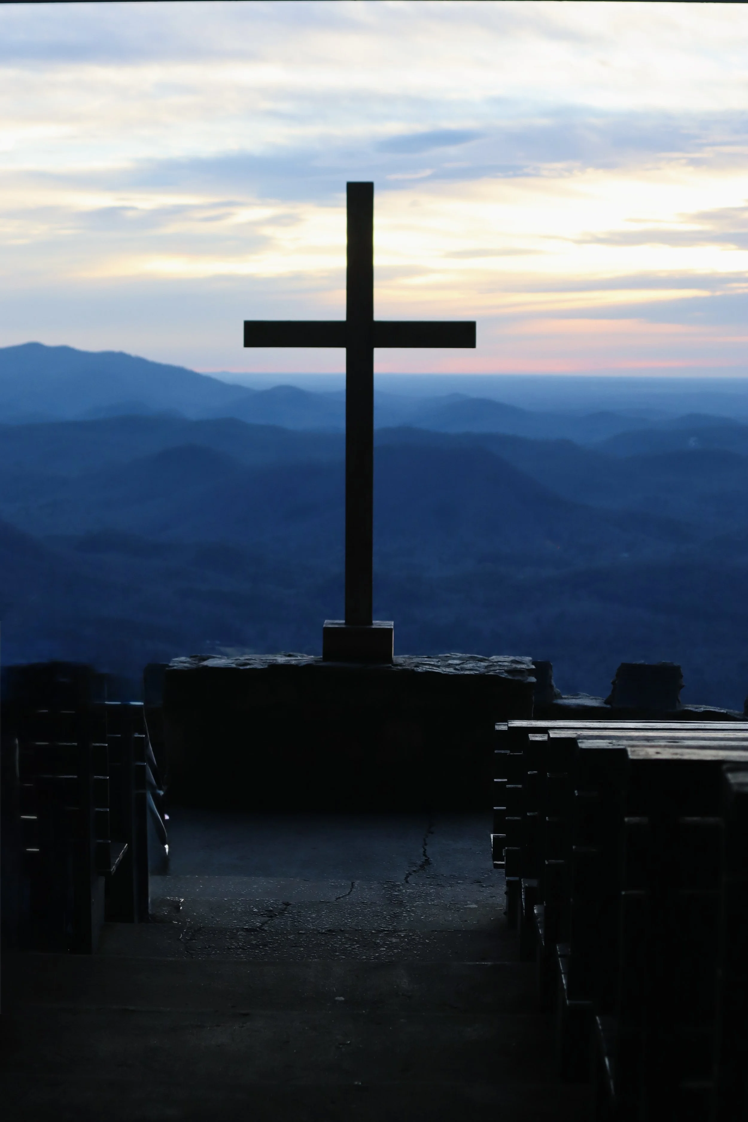 Silhouette of a Christian cross on a stone platform overlooking mountainous landscape at sunset