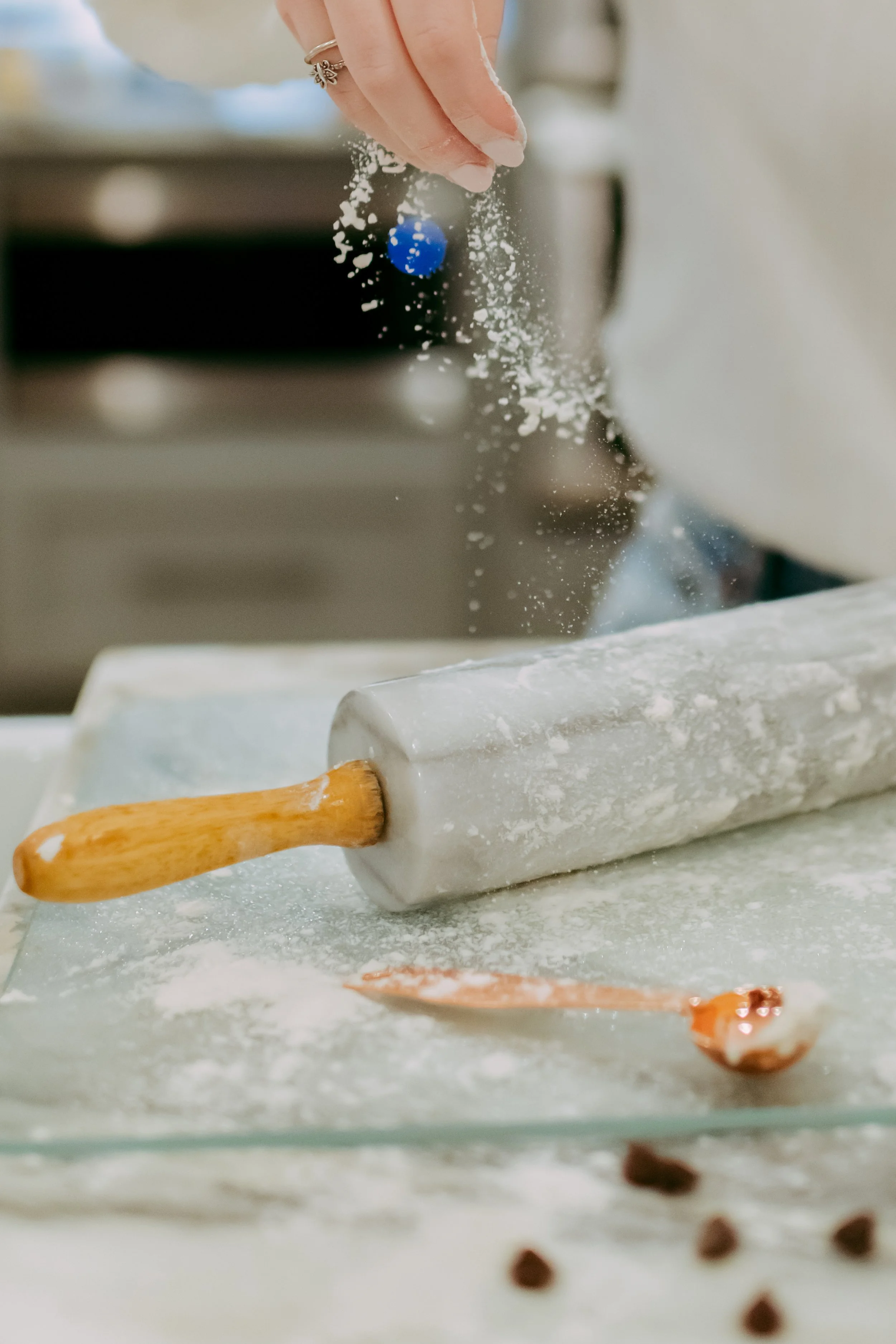 A hand dusting flour over a kitchen counter with a pastry rolling pin and a spoon nearby.