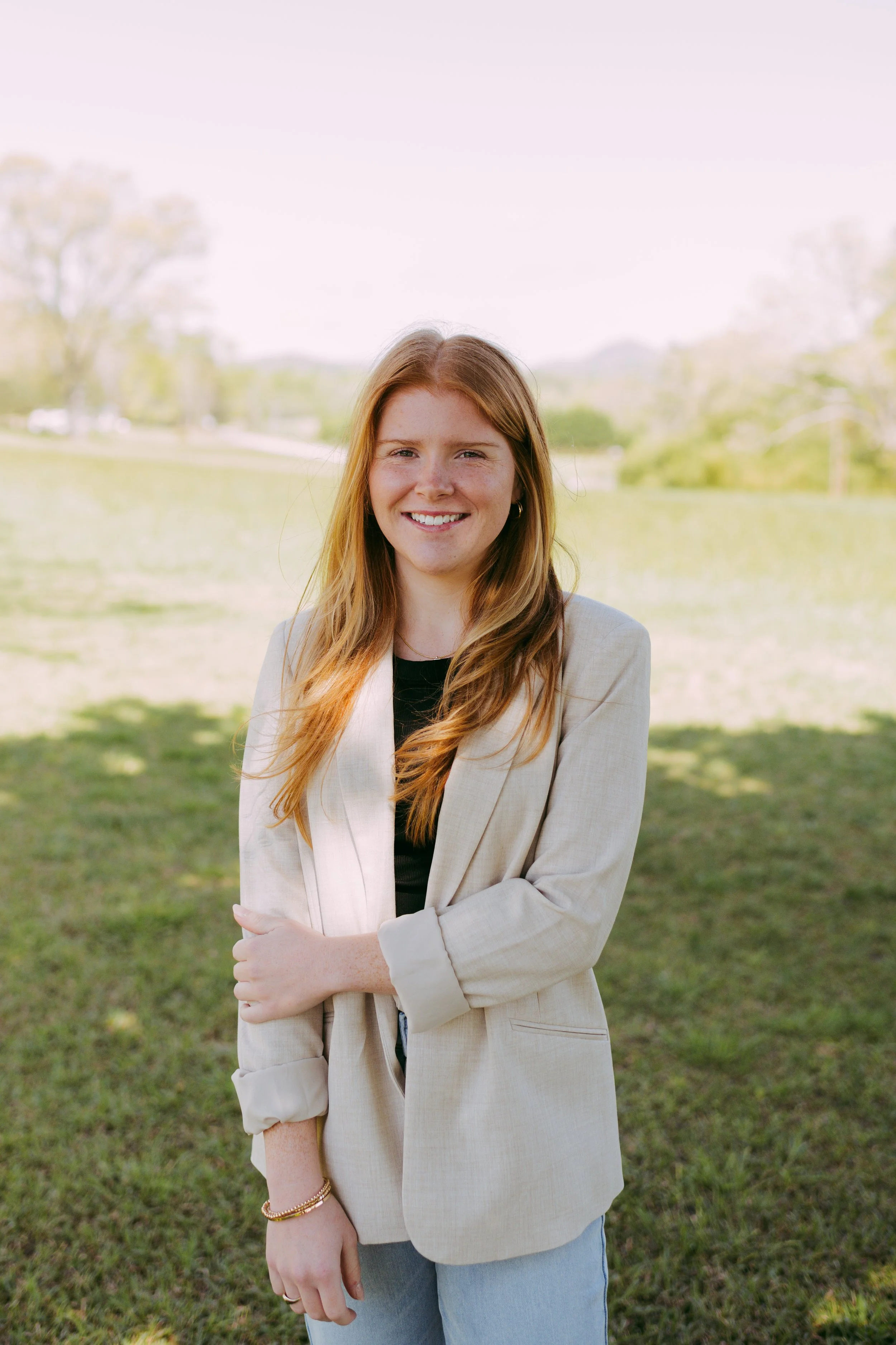 A young woman with long red hair, smiling, standing outdoors on a sunny day with a grassy field and trees in the background.