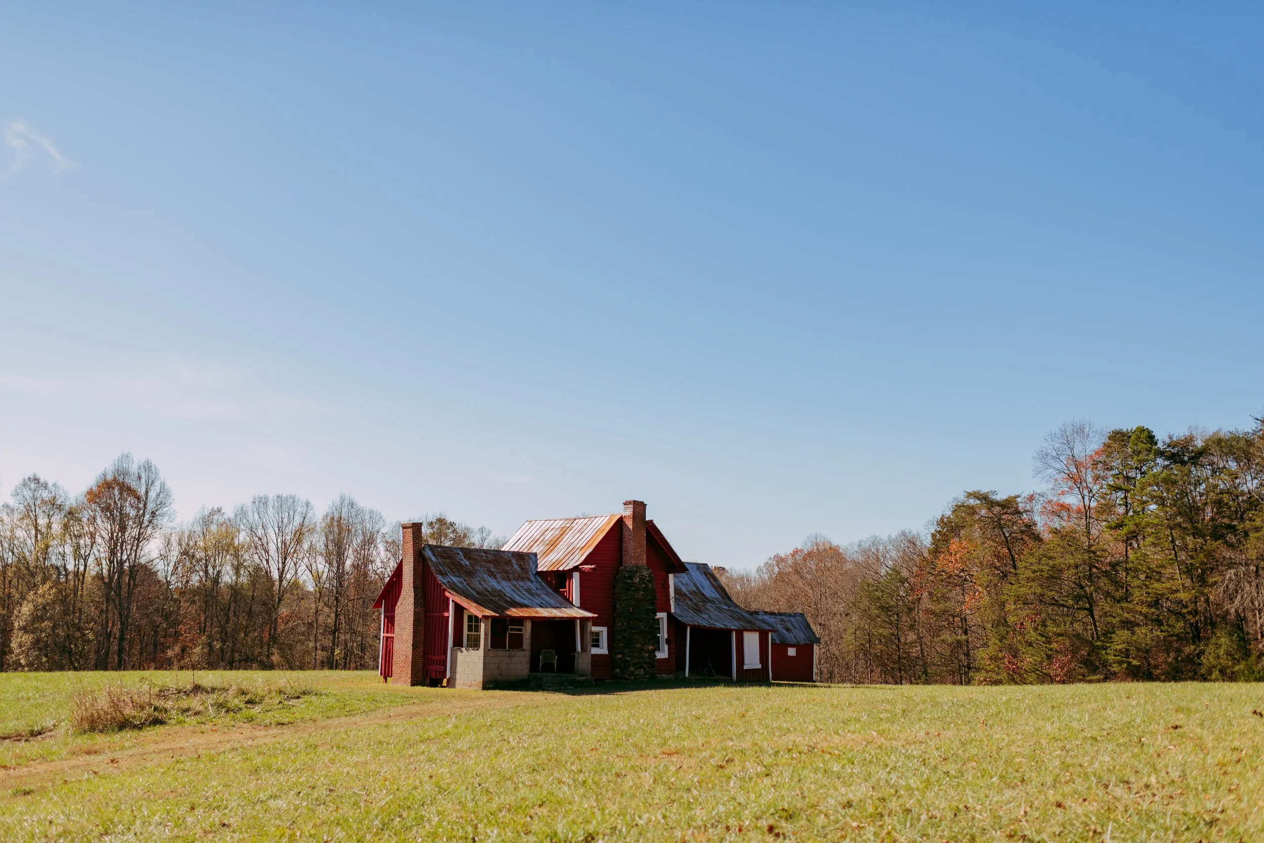 A rustic red house with a weathered metal roof situated on a grassy field, with a background of trees under a clear blue sky.
