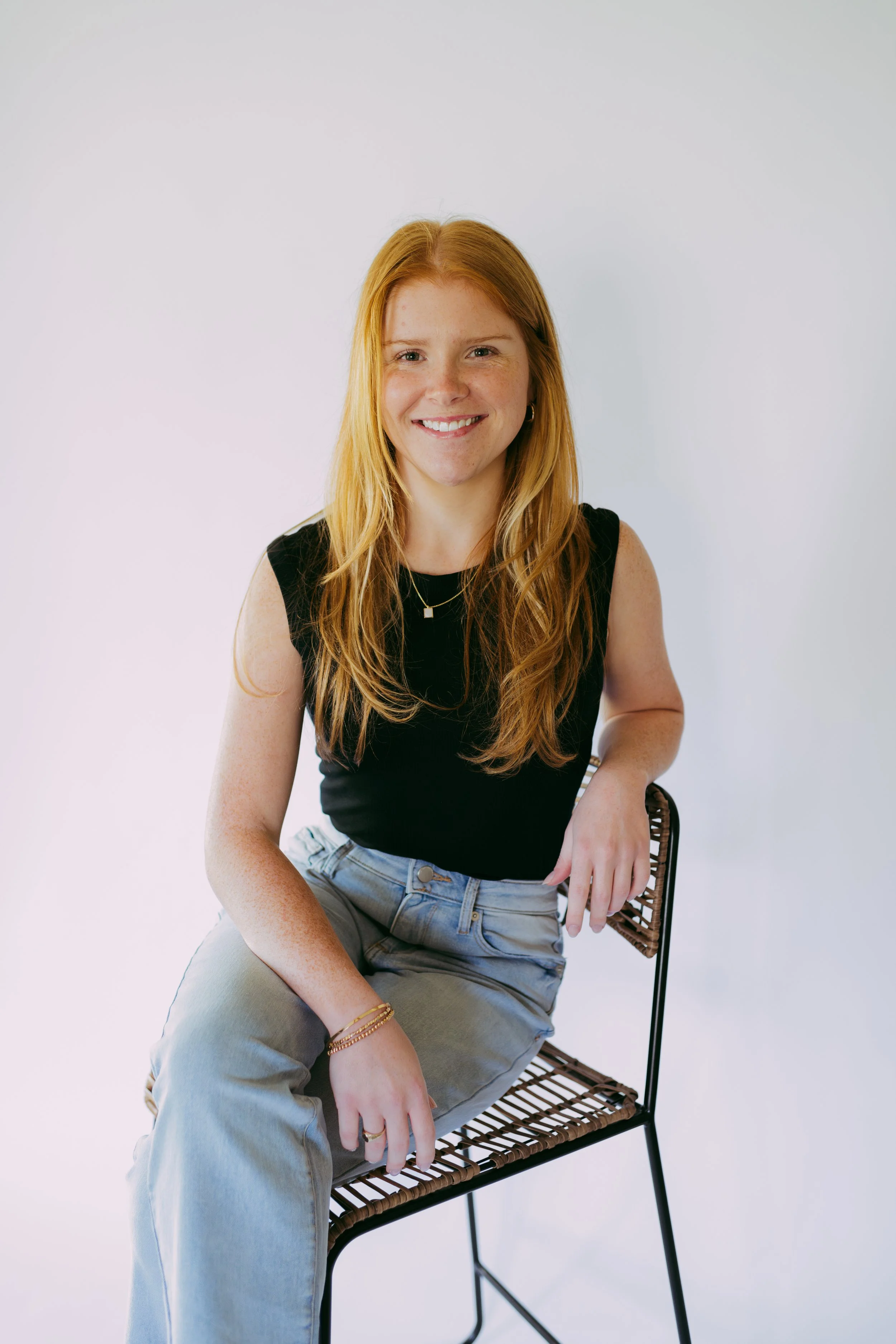 A smiling woman with long red hair wearing a black sleeveless top and light blue jeans, sitting on a modern chair with a white background.