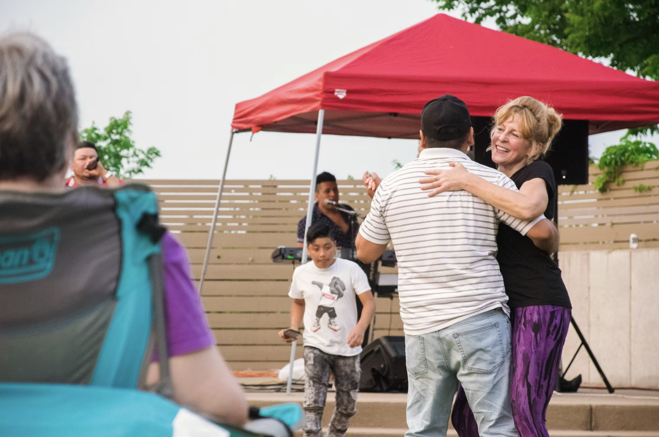 A woman and a man dancing and hugging at an outdoor event with a red canopy tent, a fence, and trees in the background. A musician is playing keyboards in the background, and a child stands nearby.
