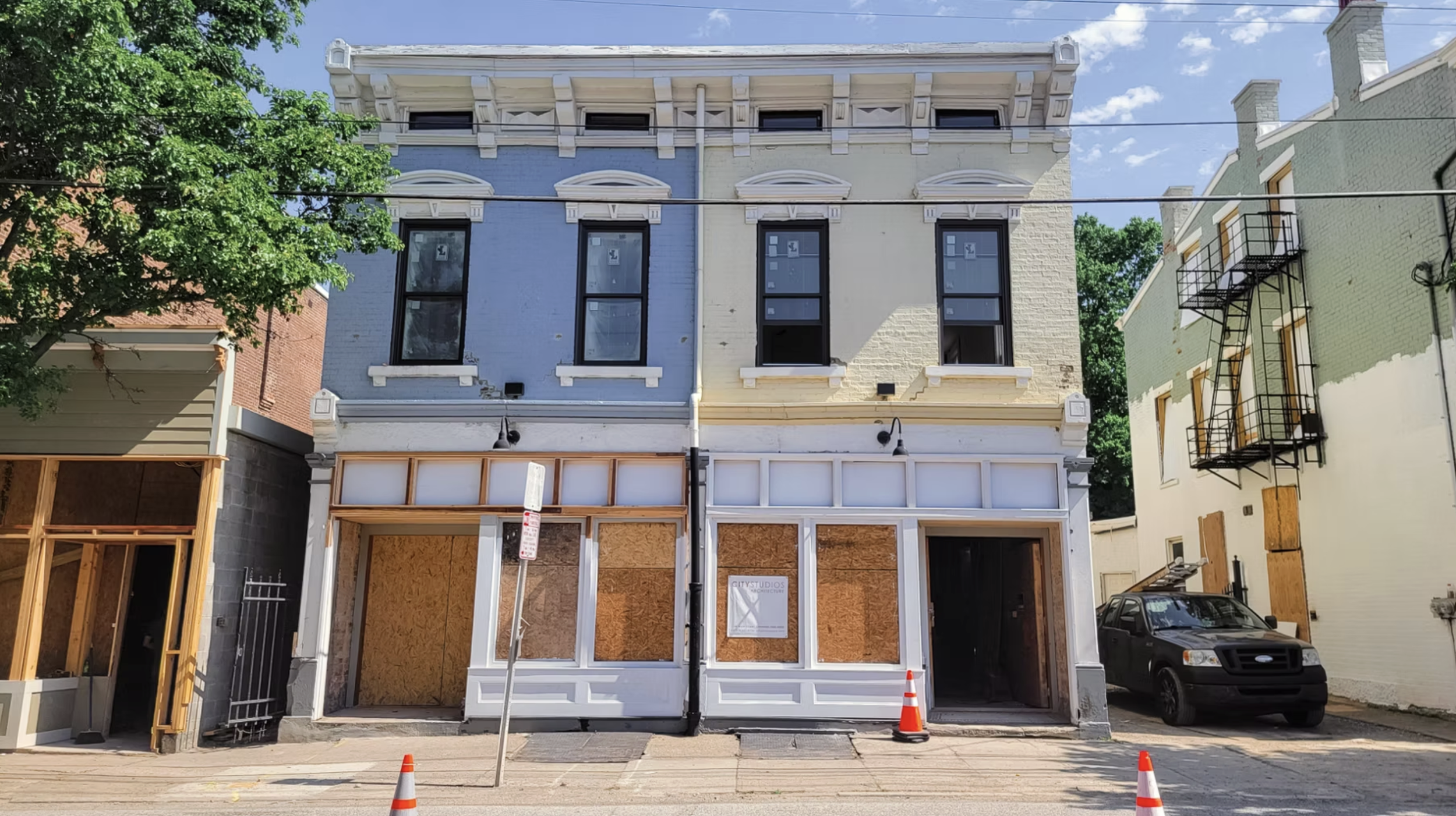 A row of three-story buildings, with the middle building under renovation, boarded up with plywood on the ground floor. The building is painted in light blue and cream, with black window frames. To the right, a black vehicle is parked on a small driv