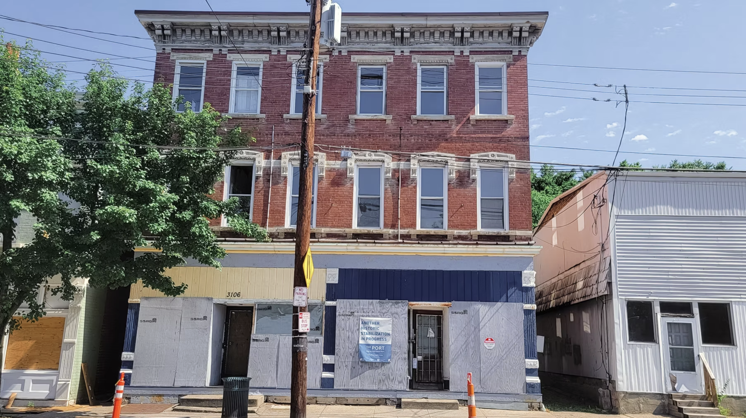 A three-story brick building under construction with boarded-up windows and a construction sign, adjacent to a white building, with a tree and utility pole in front.