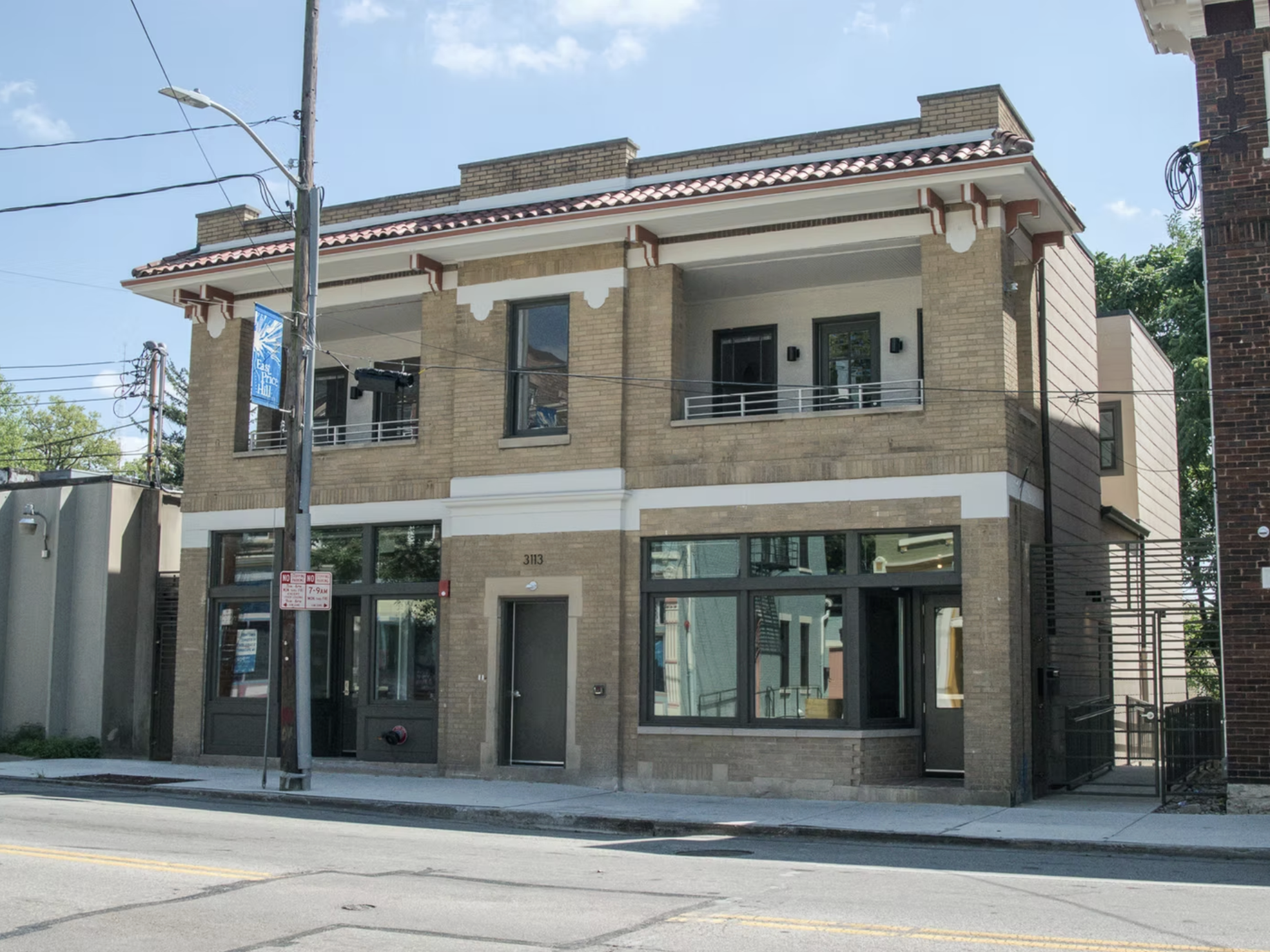 A three-story modern building made of brick and large windows, situated on a city street with utility poles and wires in front. The building has a small second-floor balcony with a metal railing.