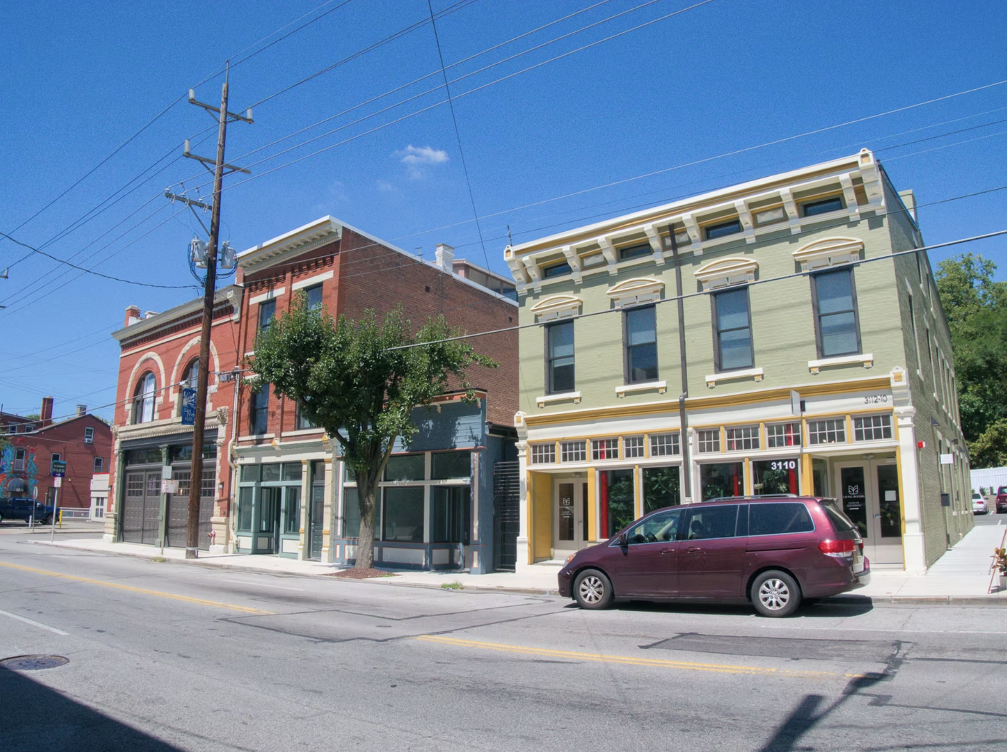 Street view of a building with a green facade and another with a red brick facade, parked maroon minivan, tree, and overhead power lines under a blue sky.