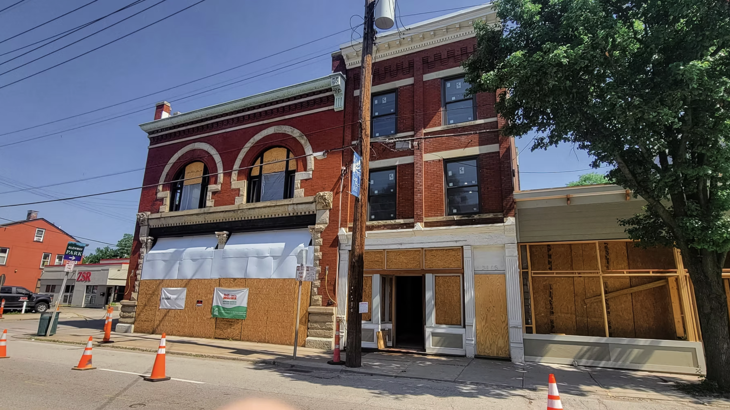 Street view of a three-story brick building under renovation with boarded-up windows and construction barriers, orange cones, and a tree in the foreground.