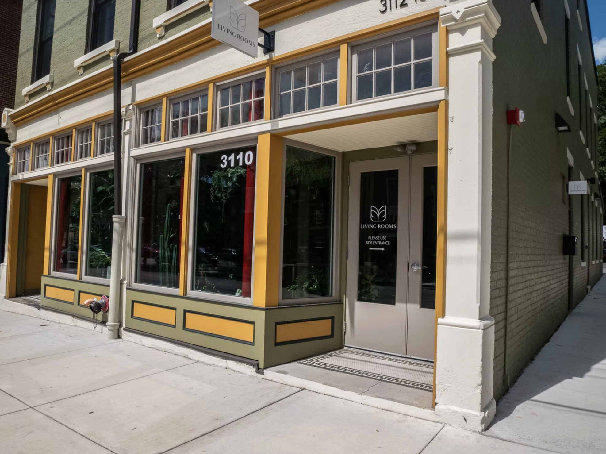 Exterior view of a building with large windows, green and yellow trim, and a glass door labeled 'Living Rooms.' The address '3110' is displayed above the door, and a sign for 'Living Rooms' is mounted above. The sidewalk is visible in front of the bu