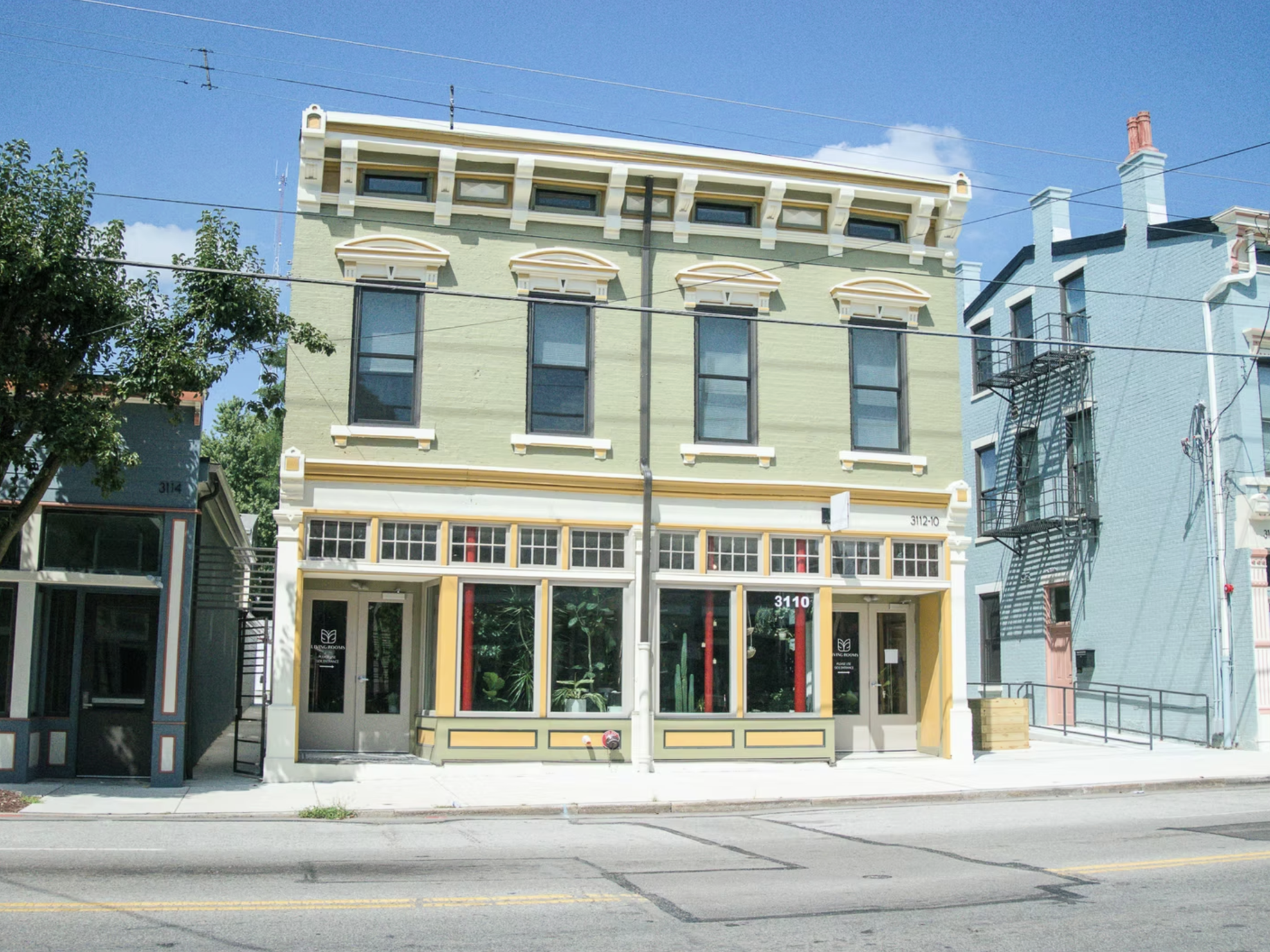 Colorful three-story building on a sunny street, with large windows on the upper floors and storefront windows with plants on the ground floor.